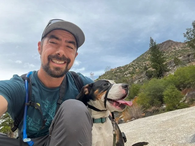 A man smiling with a dog outdoors in a mountain setting.