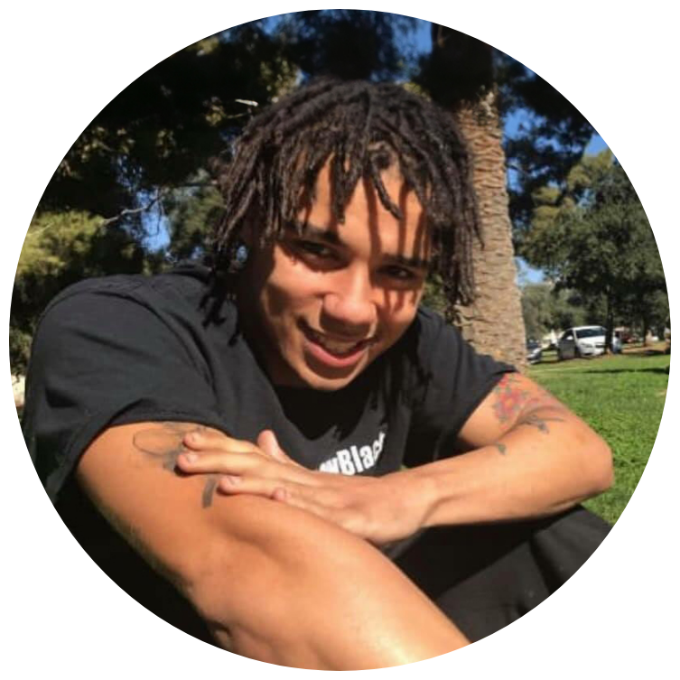 Young man with dreadlocks sitting outside on grass, smiling, with trees and cars in the background.