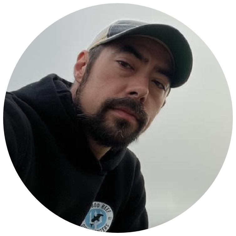 A man with a beard and mustache wearing a black jacket and a camo cap, outdoors on a cloudy day.