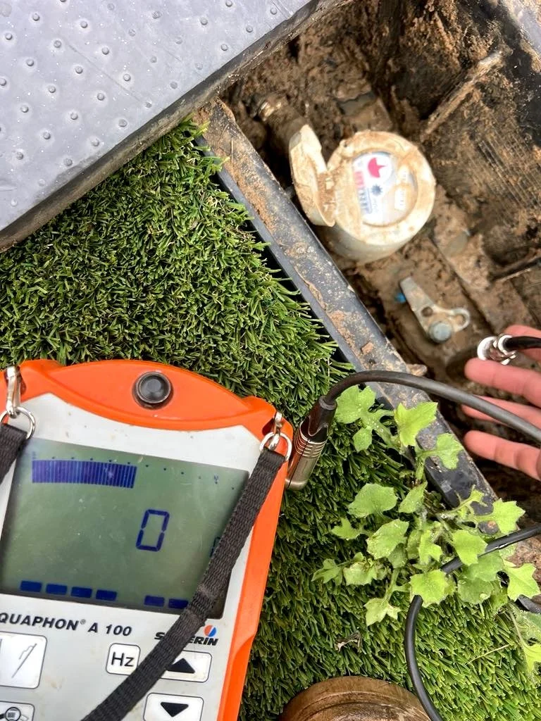 Close-up of a soil testing device measuring the soil near green plants, with the soil probe inserted in the ground, surrounded by environmental elements including greenery and a hand holding electrical testing cables.