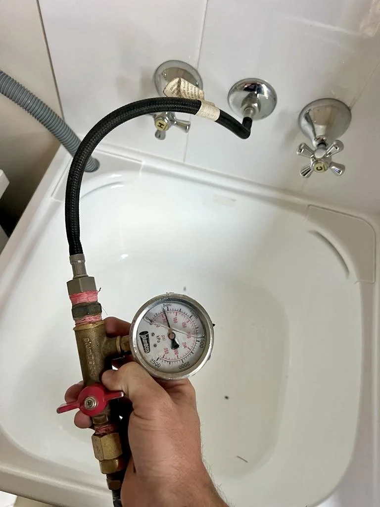 Person holding a pressure gauge connected to a hose over a white sink with plumbing fixtures in the background.