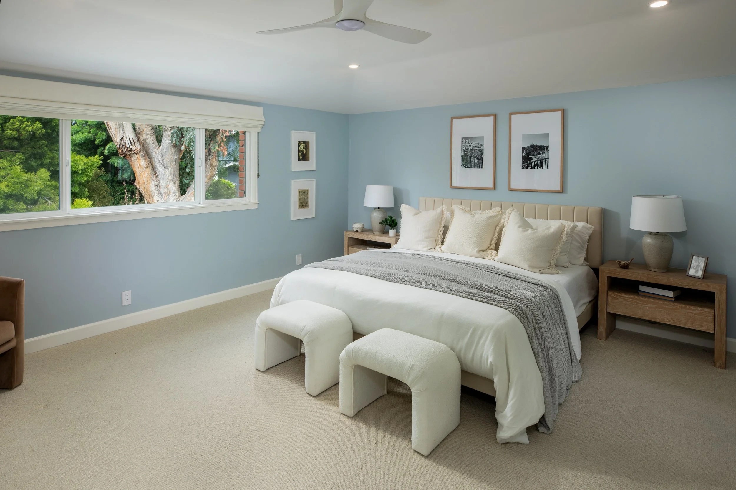 A bedroom with a large window showing trees outside, light blue walls, a bed with white and beige bedding, a wooden nightstand with a lamp and potted plant, and framed artwork on the wall.