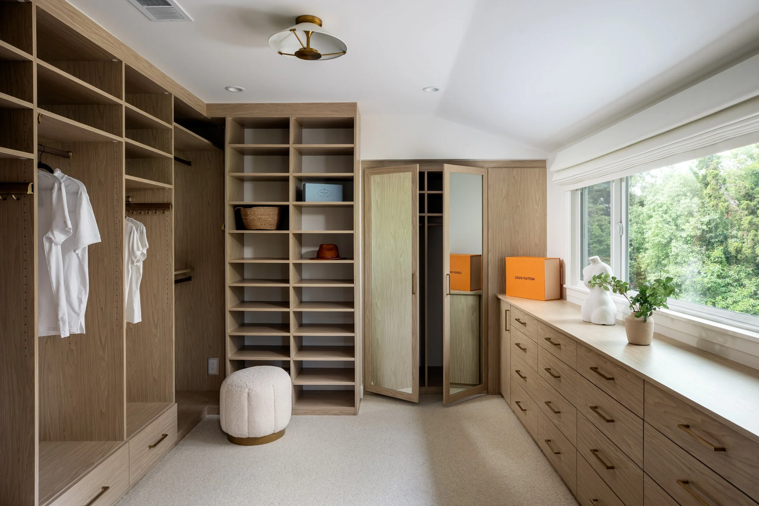 A walk-in closet with wooden shelving and drawers, a large window with greenery outside, and decorative vases on the window sill.