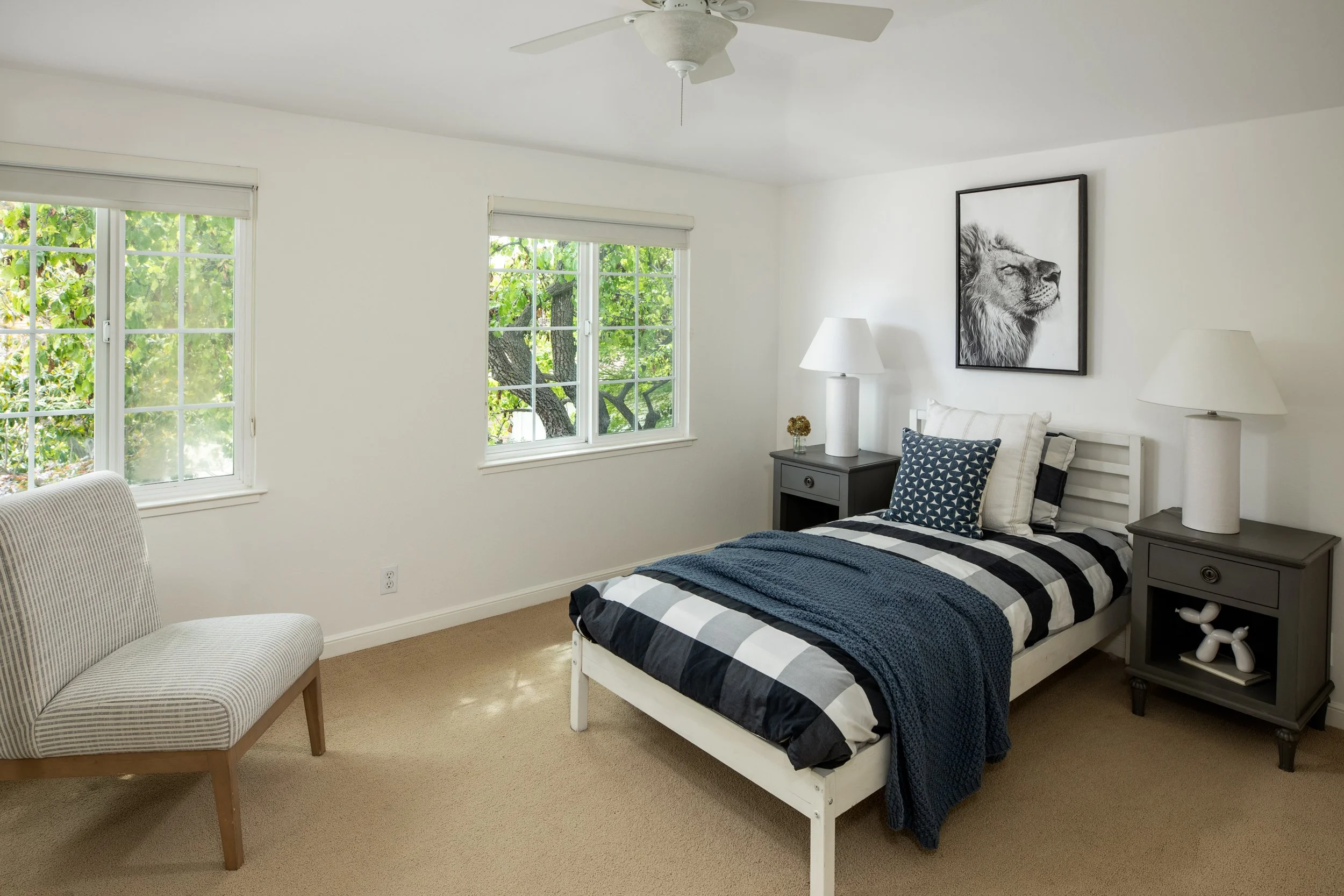 A bedroom with two windows, a white bed with black and white striped bedding, a dark gray nightstand with a lamp and decorative items, a wall art of a lion, a white ceiling fan, and a beige carpeted floor.