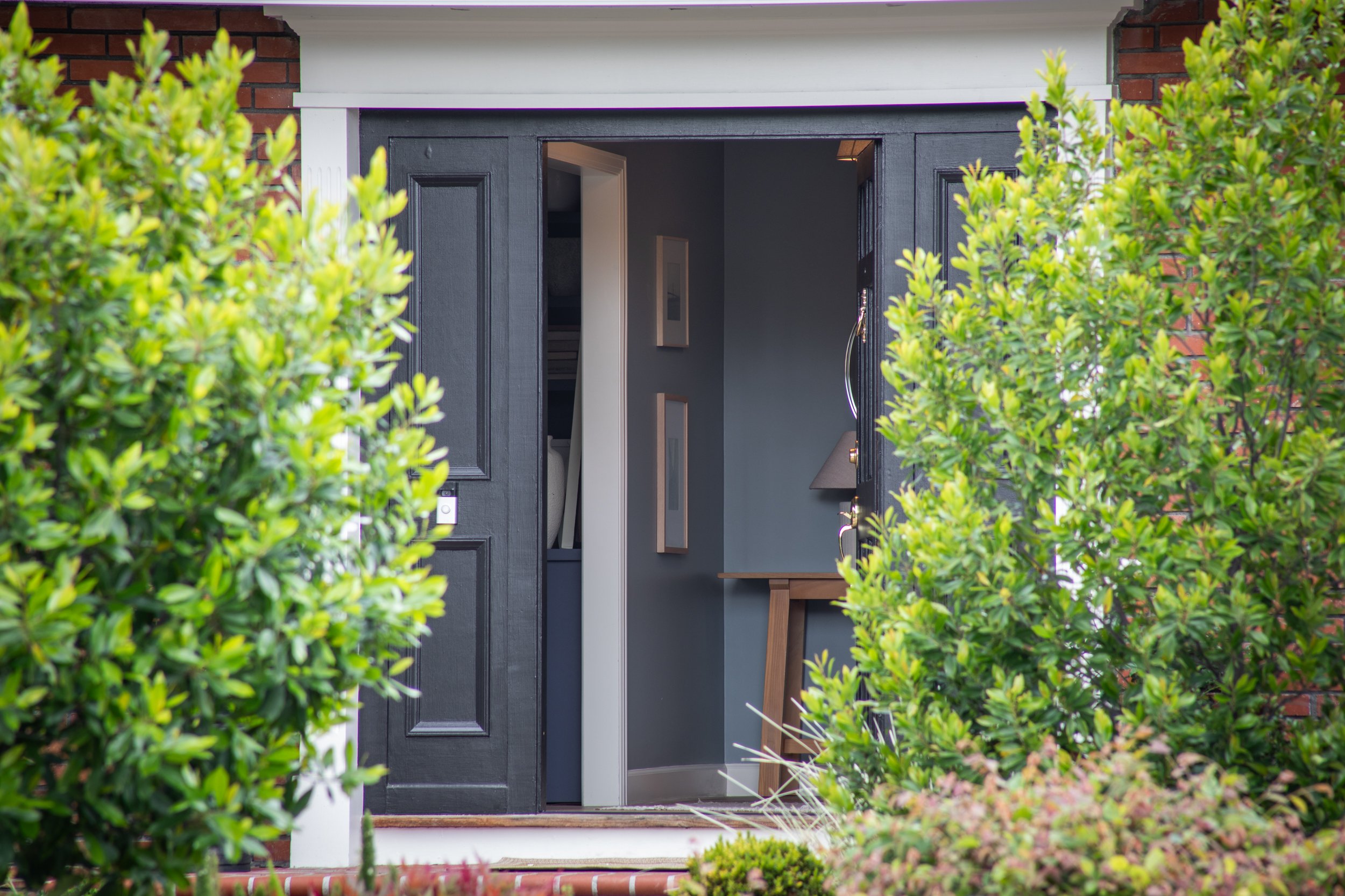 Open black front door with interior visible, framed by lush green bushes on both sides, brick exterior visible on the sides, leading into a home with a doorway and interior furnishings