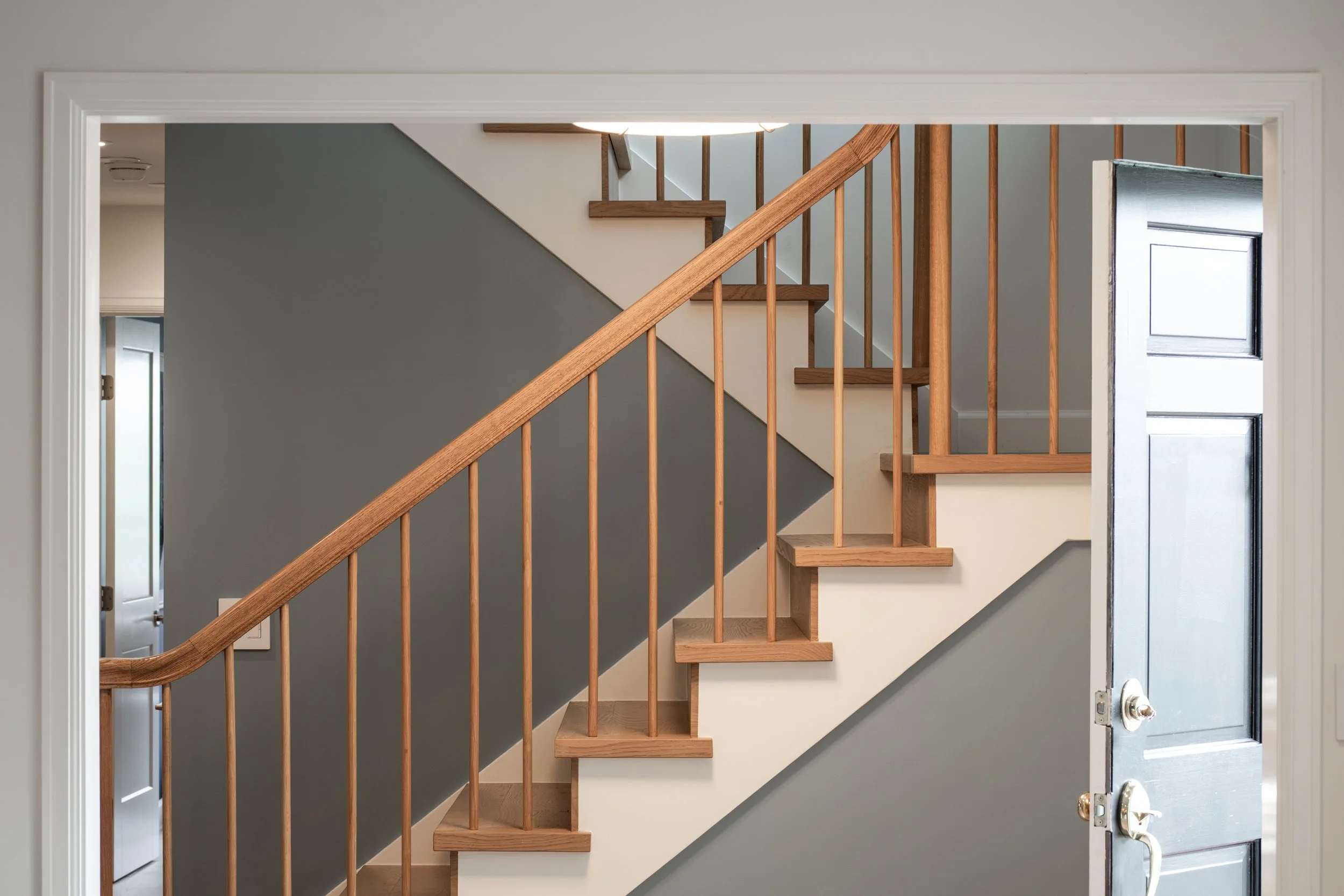 Interior view of a staircase with wooden handrail and balusters, beige stairs, a gray accent wall, and a half-open door to the left, with a window on the right side near the door.