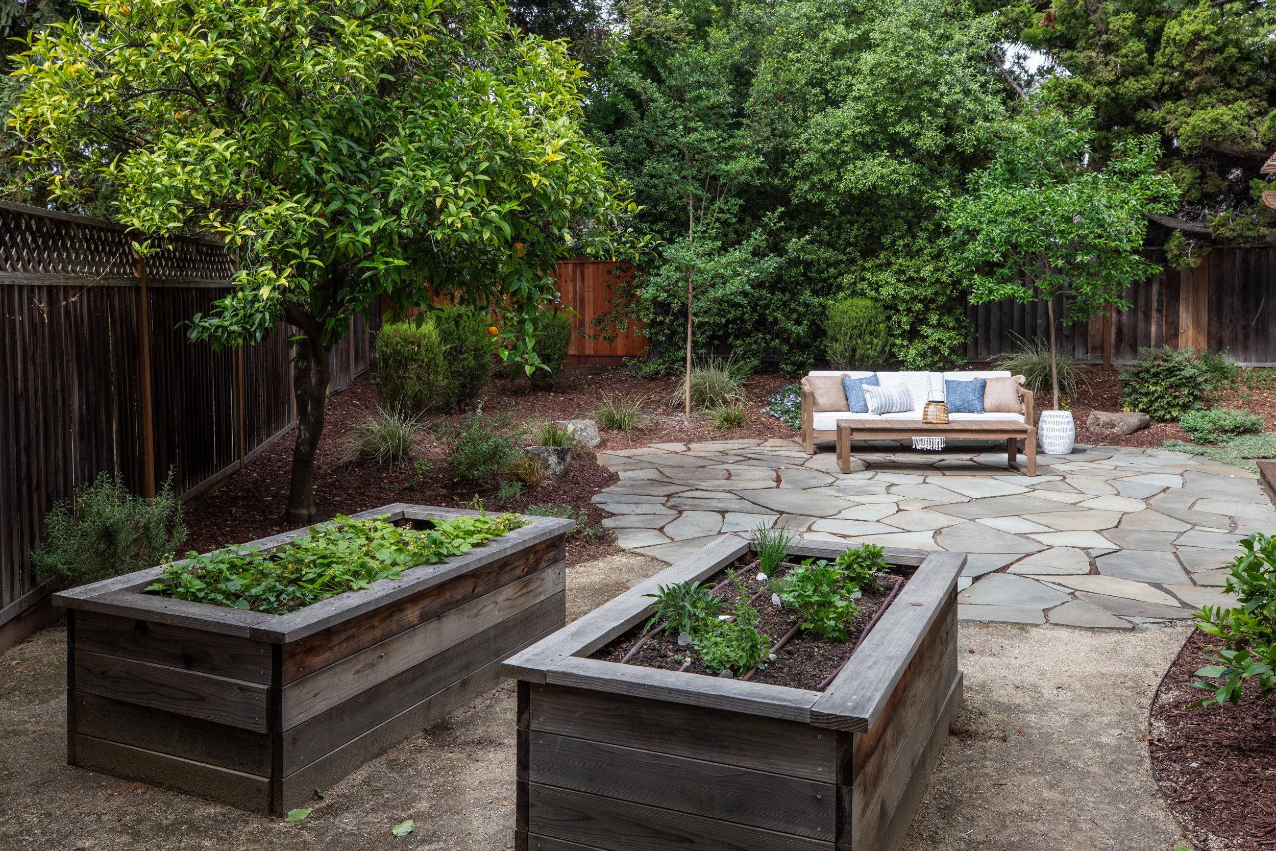 A backyard garden with a stone patio, a white outdoor sofa with blue and beige pillows, surrounded by lush green trees and plants, and a wooden fence in the background.