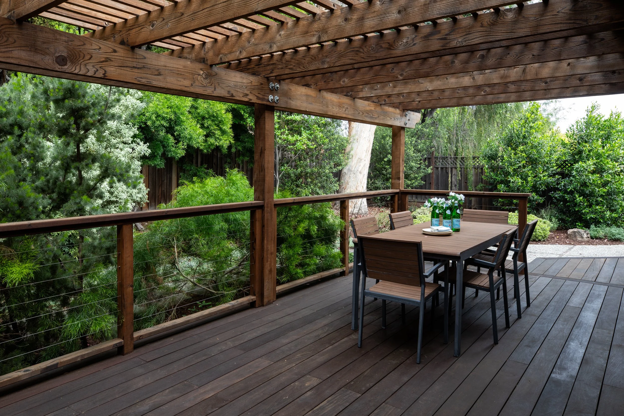 Outdoor wooden deck with dining table, chairs, greenery, and a pergola overhead.