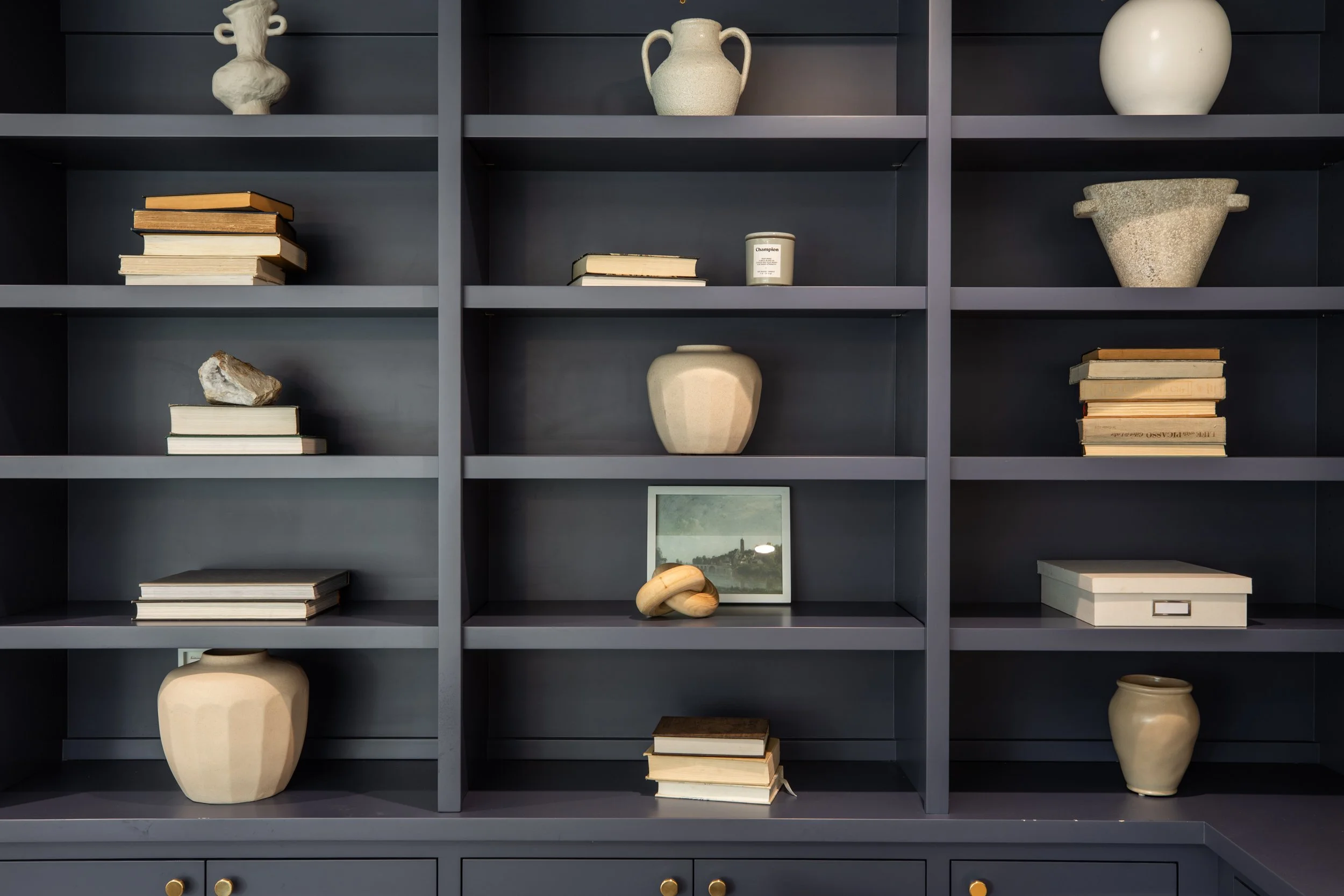 A bookshelf painted in dark gray with multiple compartments. The shelves hold an assortment of books, pottery, and decorative objects, including vases, a framed picture, and a stone sculpture.
