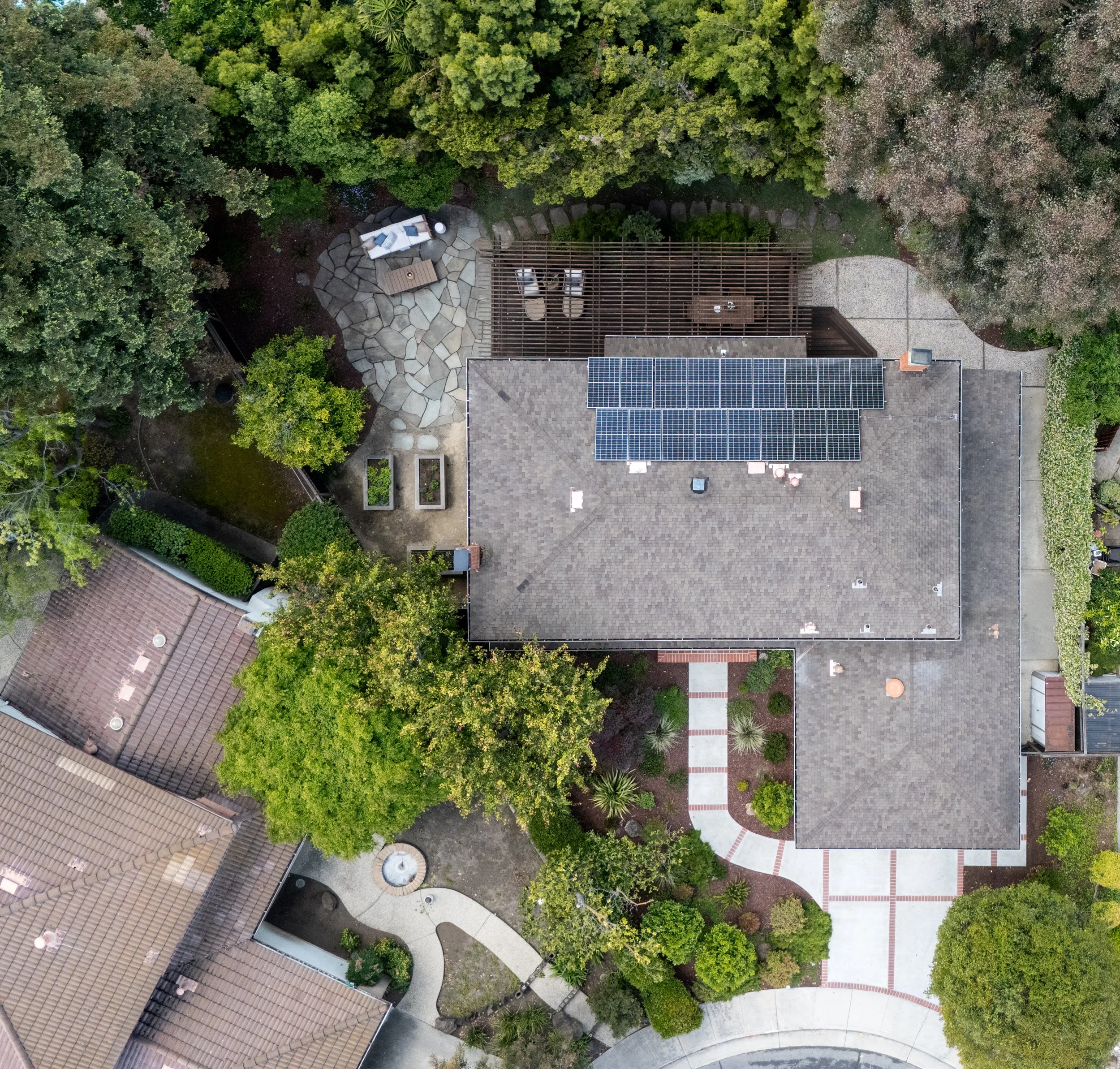 An aerial view of a backyard with a patio area featuring outdoor furniture, surrounding lush green trees, and part of a house with solar panels on the roof.