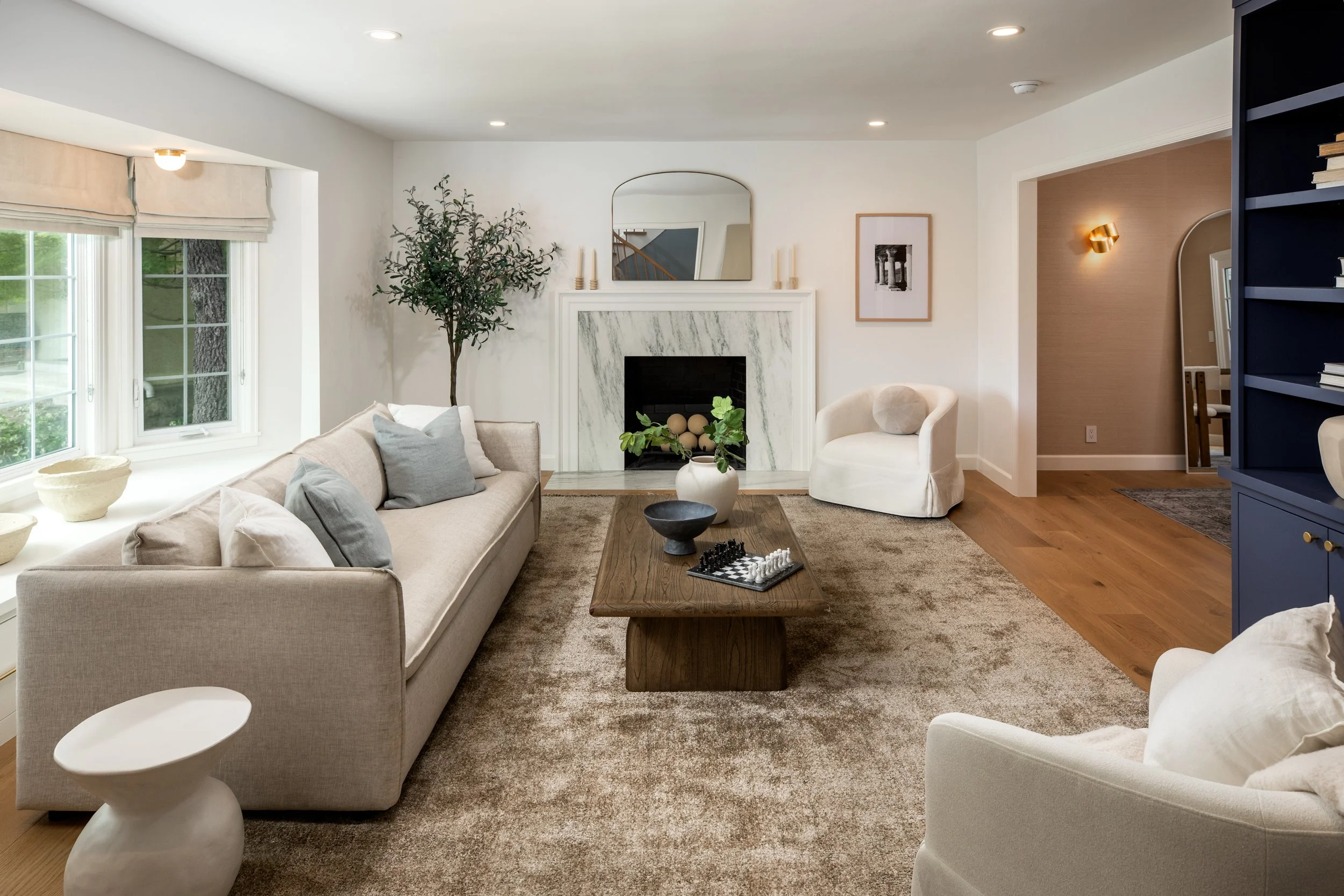 Living room with beige sofa, white armchair, fireplace with marble surround, wooden coffee table with chess set, potted plant, and large window with blinds.