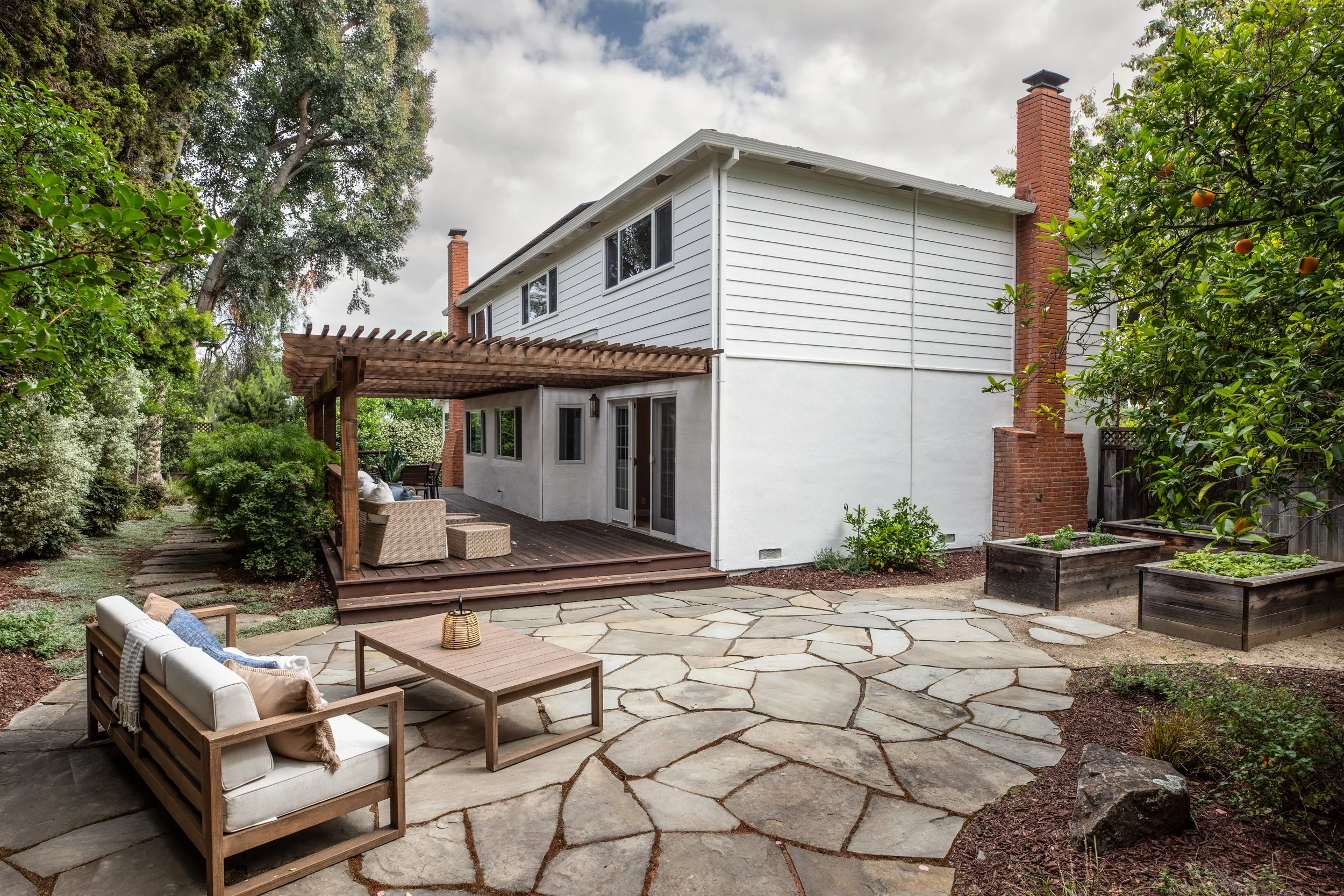 A backyard patio area with stone paving, outdoor seating, a wooden pergola, lush greenery, and a white two-story house with brick chimneys in the background.