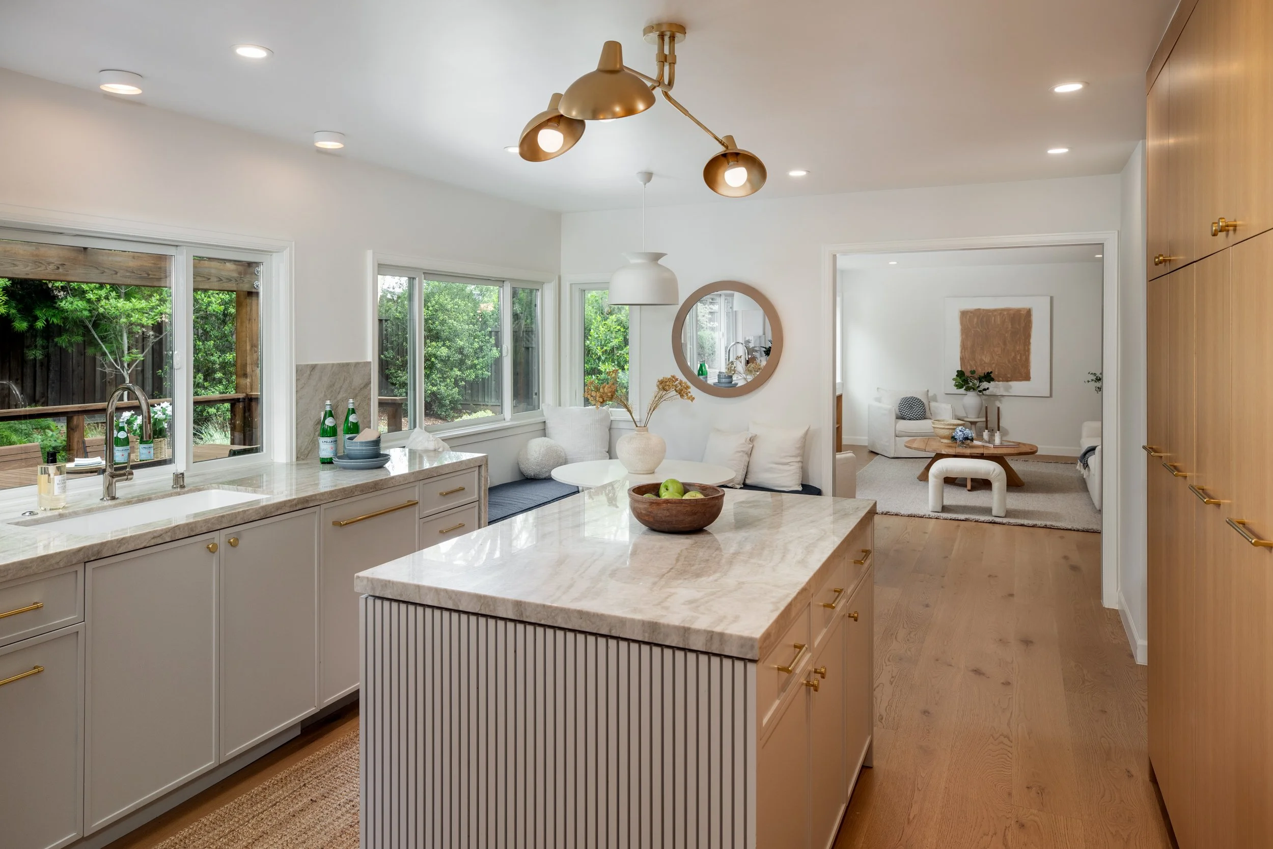 Modern kitchen with white cabinets, marble countertops, and a large island. Open living space with windows looking out to a backyard with greenery. Decor includes a round mirror, vases with dried flowers, and simple furniture.