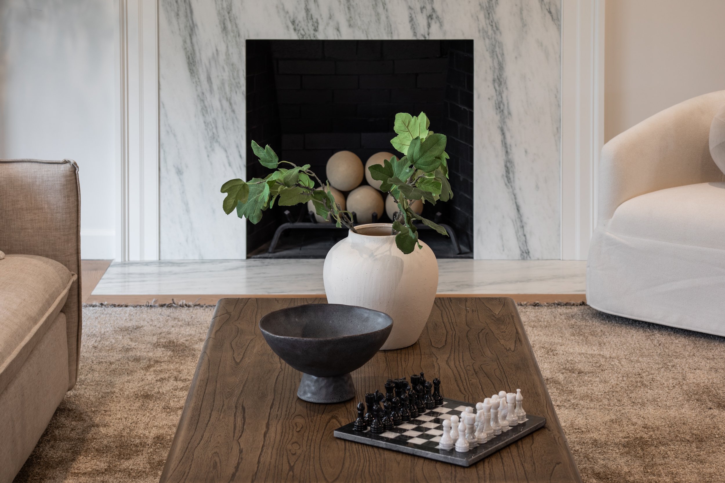 Living room with a marble fireplace, a potted plant, a chessboard on a wooden coffee table, and beige furniture.
