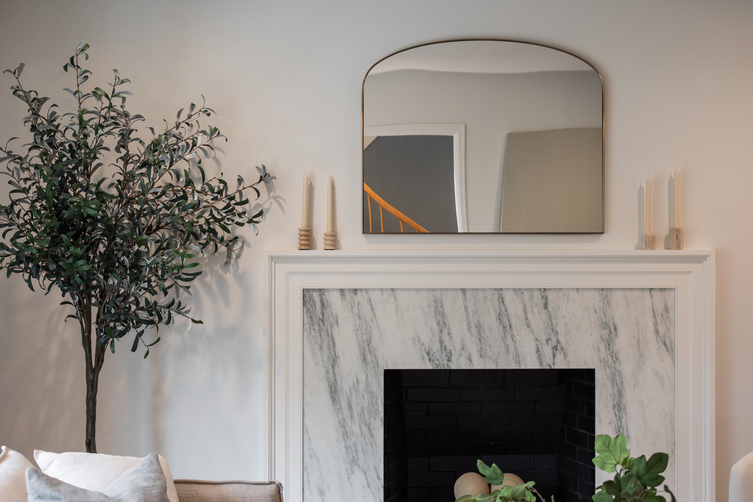 Living room with a white fireplace, marble surround, wood mantel, and a large mirror above. To the left, a tall leafy green plant and the edge of a sofa with pillows are visible.