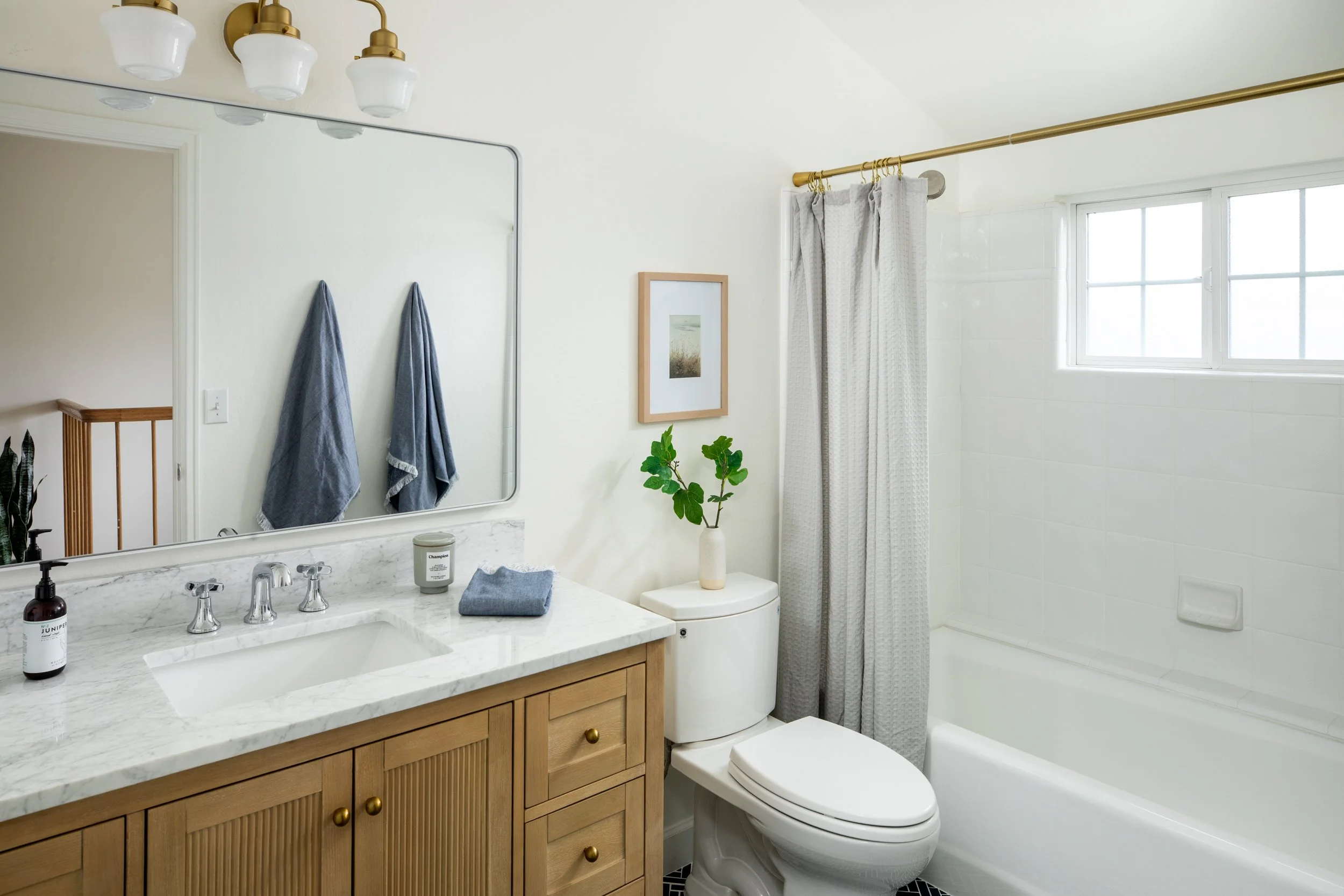 A modern bathroom with a wooden vanity, marble countertop, and silver fixtures. There are two hand towels hanging on the wall, a framed picture above a potted plant, and a window with white curtains over a bathtub.