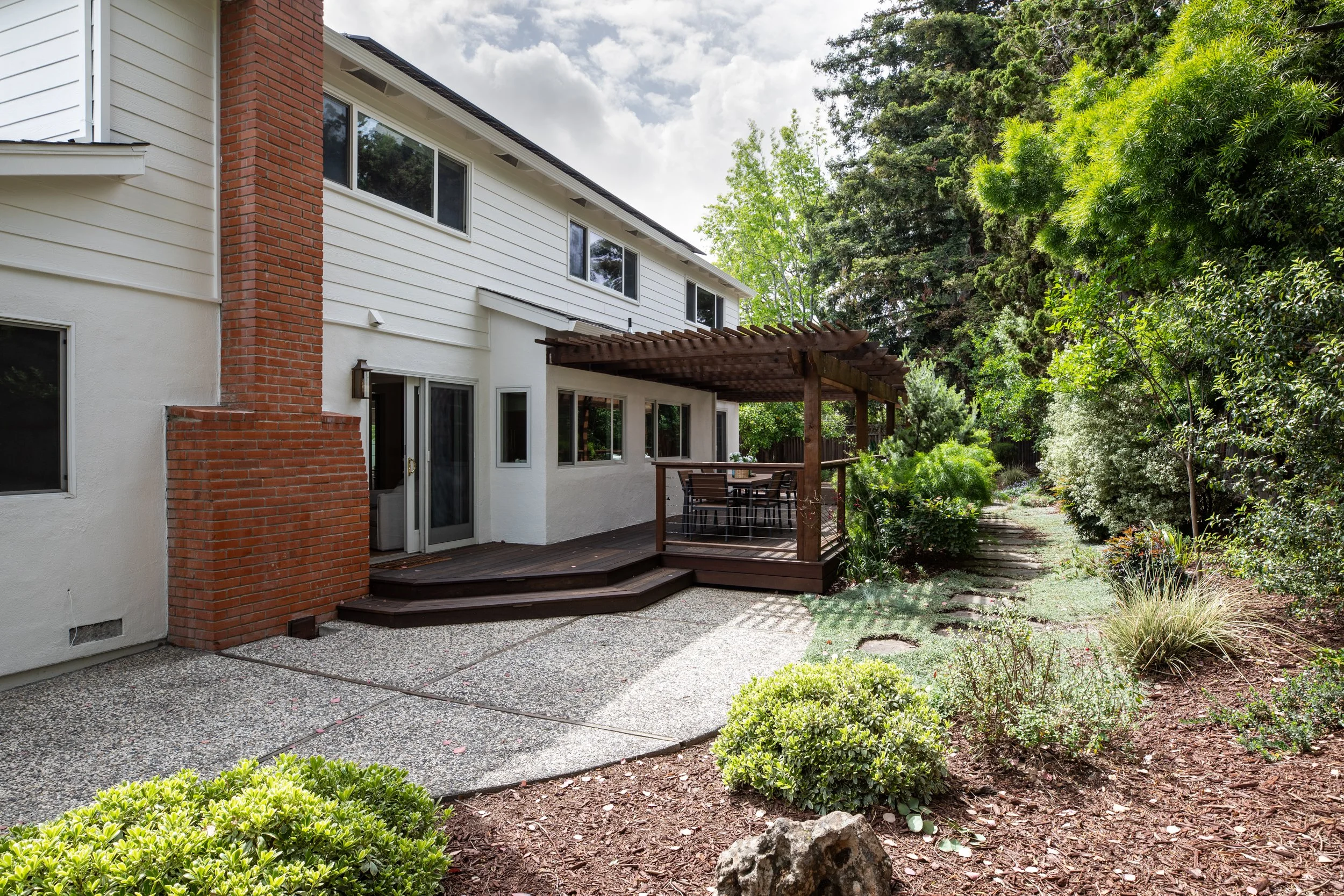 Backyard view of a white house with a red brick chimney, sliding glass doors, and a wooden patio with a table and chairs, surrounded by lush green trees and shrubs.
