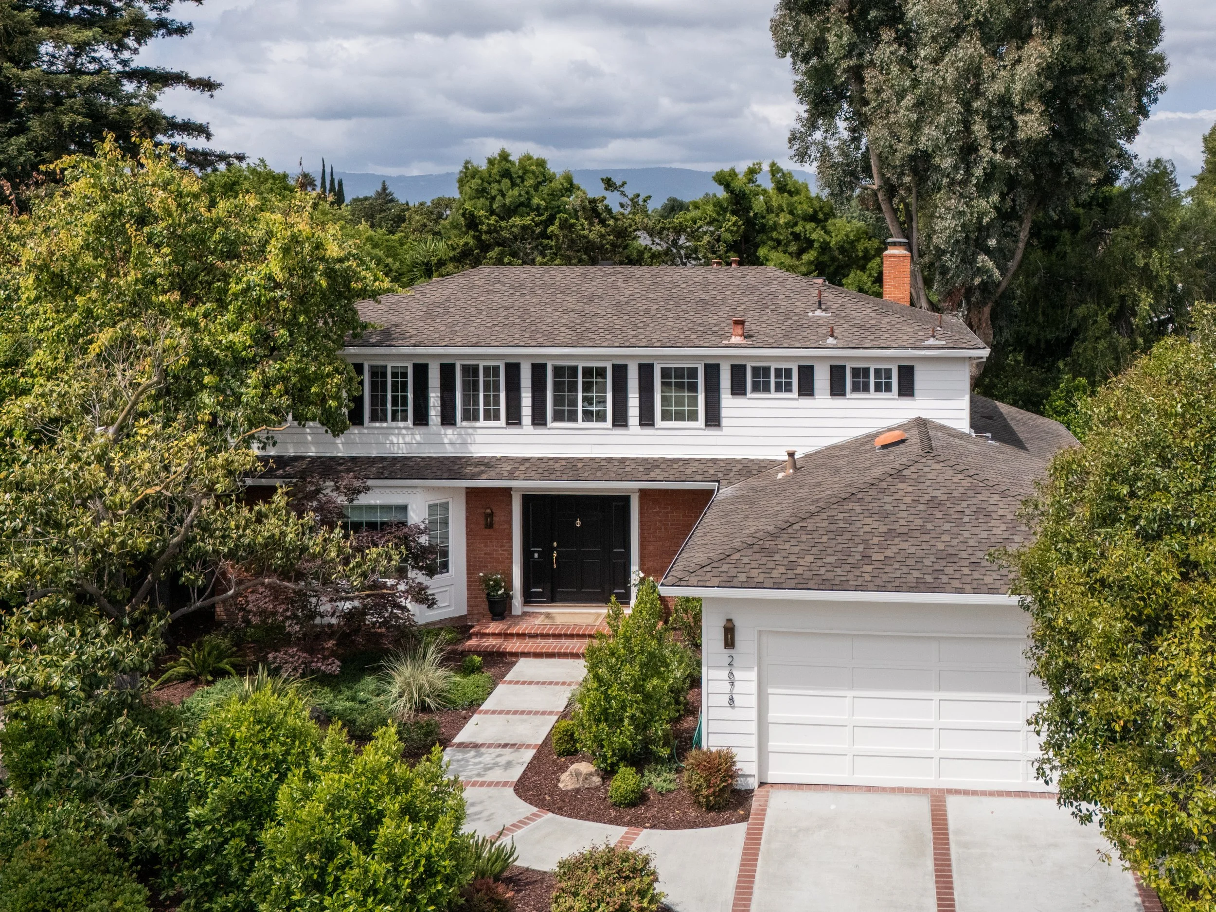 A view of a residential house with a shingled roof, white siding, black shutters, and a brick chimney, surrounded by lush greenery and trees, with a cloudy sky in the background.
