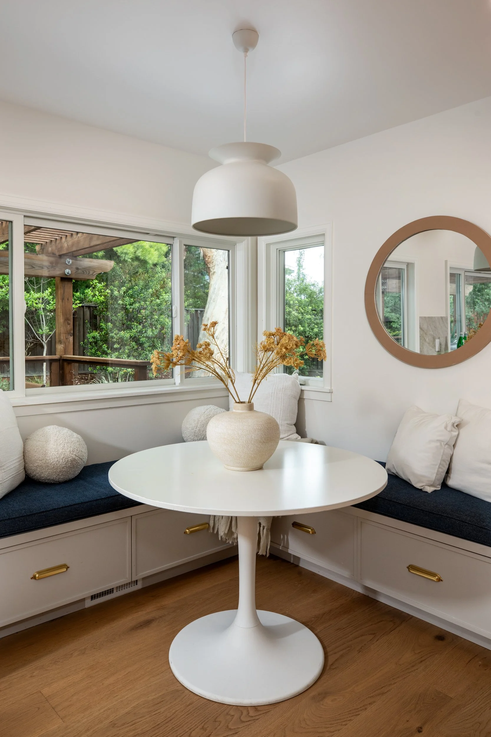 A cozy interior corner with a round white table, a built-in white bench with a cushion, pillows, and a vase with dried flowers. A large circular mirror hangs on the white wall, and sunlight shines through windows showing greenery outside. A white han
