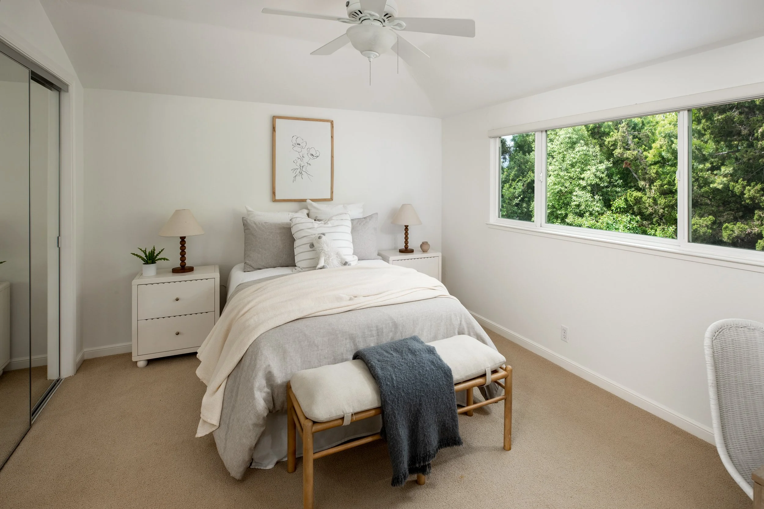 A bedroom with white walls, a bed with beige and white bedding, white pillows, and a decorative pillow with black stripes. Two white nightstands with lamps, a small potted plant, and a ceramic vase sit beside the bed. A rectangular framed line drawin