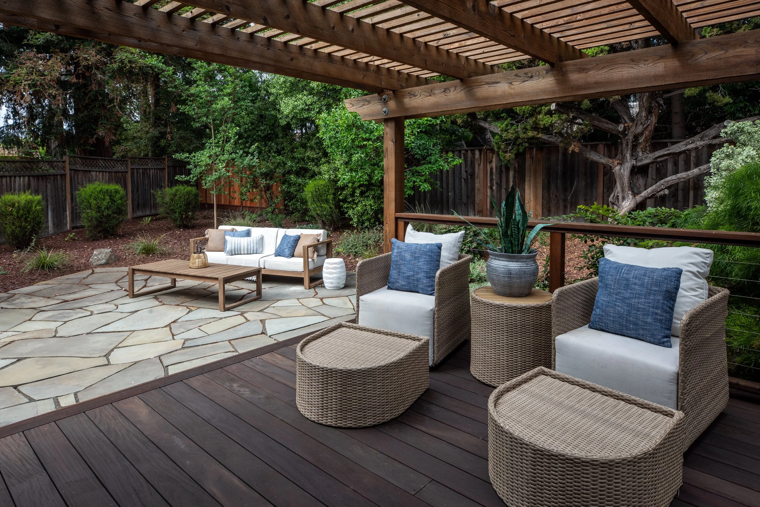 A backyard patio with outdoor furniture, including wicker chairs with cushions, a wooden coffee table, and a sofa with pillows. The patio is partially covered by a wooden pergola, surrounded by lush greenery and trees, with a wooden fence in the back