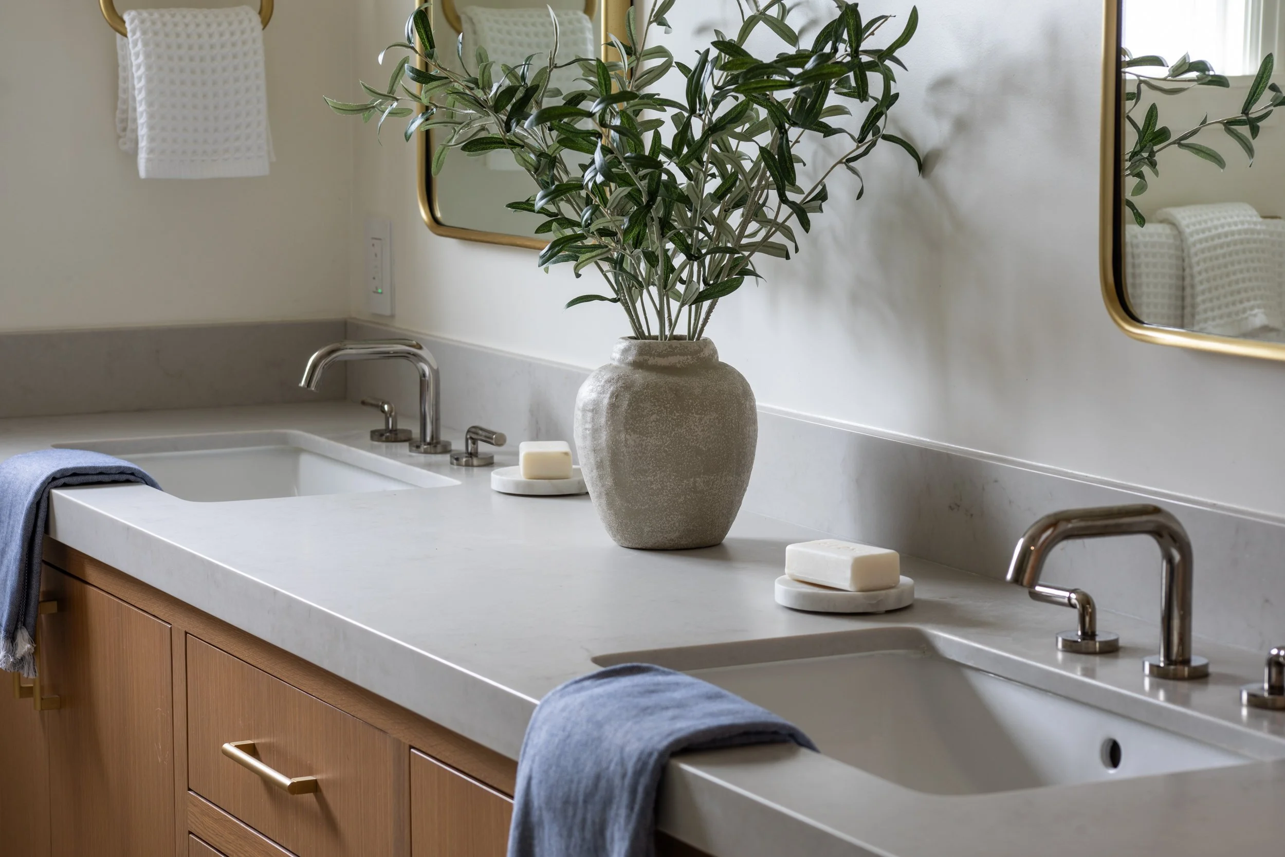 A modern bathroom countertop with a large potted plant, two soap bars, and a towel hanging over a wooden cabinet with gold handle, reflected in a mirror.