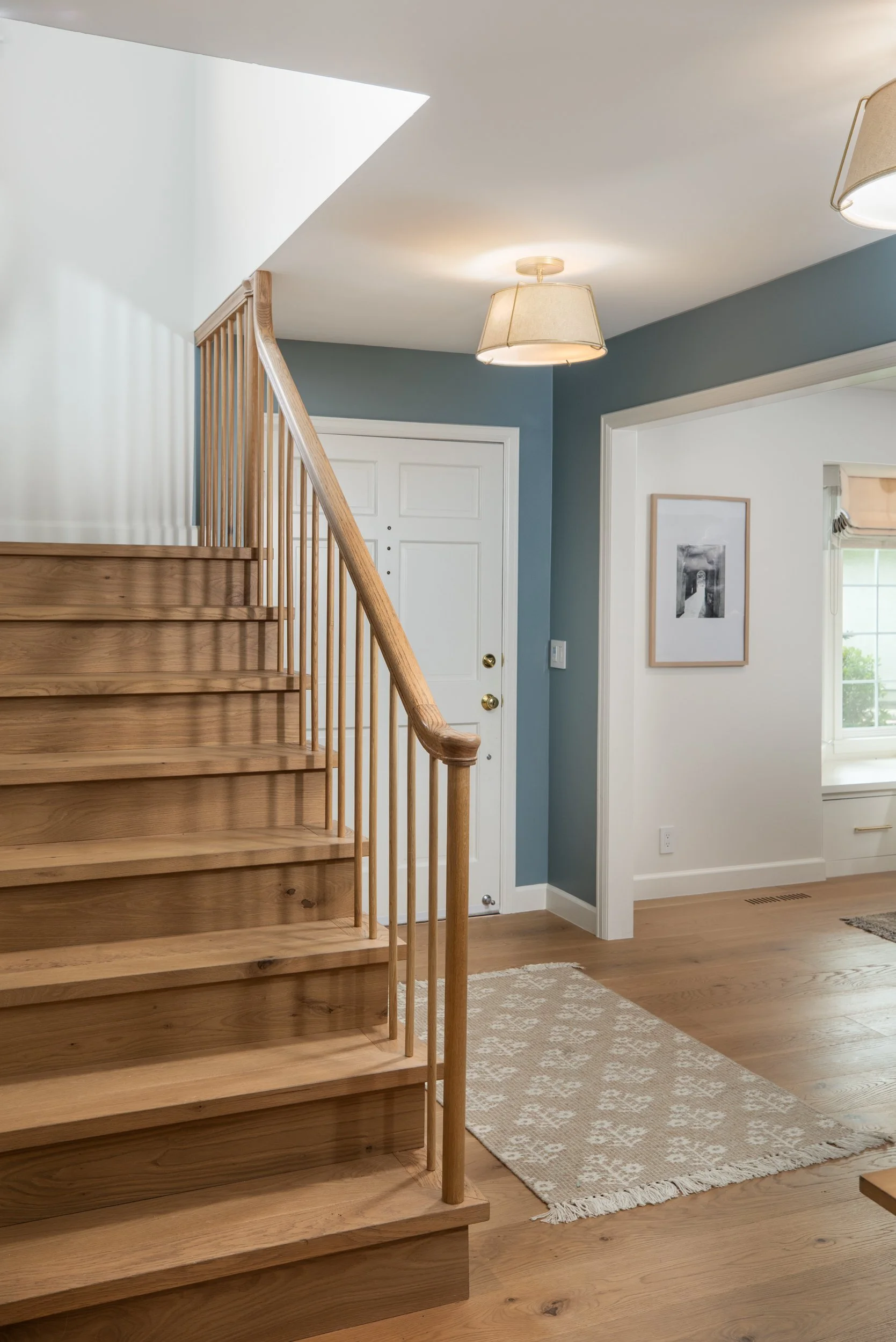 Interior of a home with wooden stairs, a white door, a framed black and white photo on a white wall, a patterned area rug, hardwood floors, and ceiling lights. The walls are painted in blue and white.