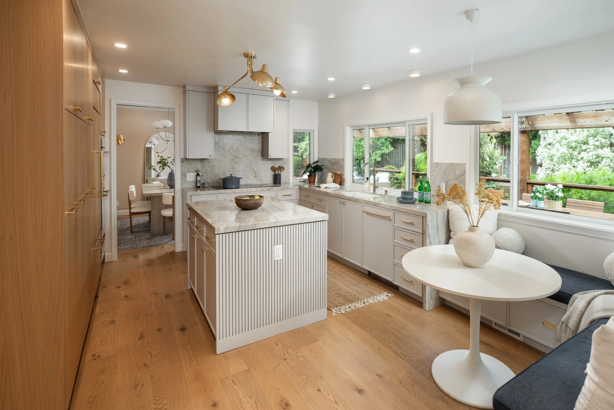 A bright, modern kitchen with white cabinets, a marble countertop, and a window overlooking greenery. There are decorative items, plants, and bottles on the counter, with a wooden dining table and a cozy seating area nearby. The space features recess