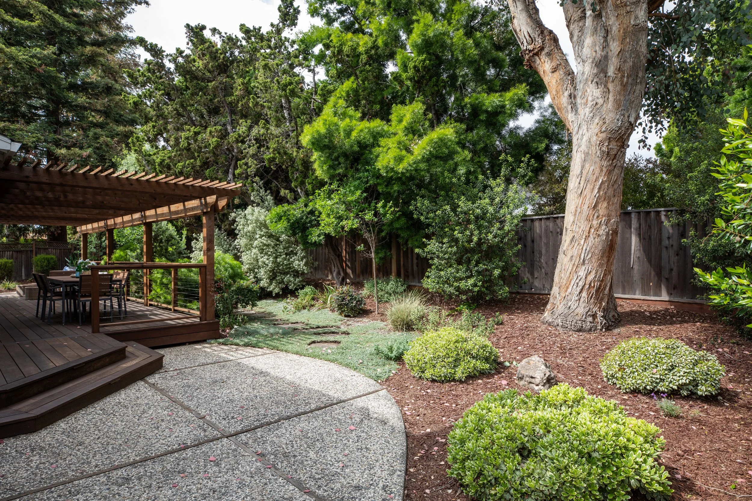 Backyard garden featuring a wooden decked dining area with a table and chairs, surrounded by lush green trees and plants, including a large tree with a textured trunk, and enclosed by a wooden fence.