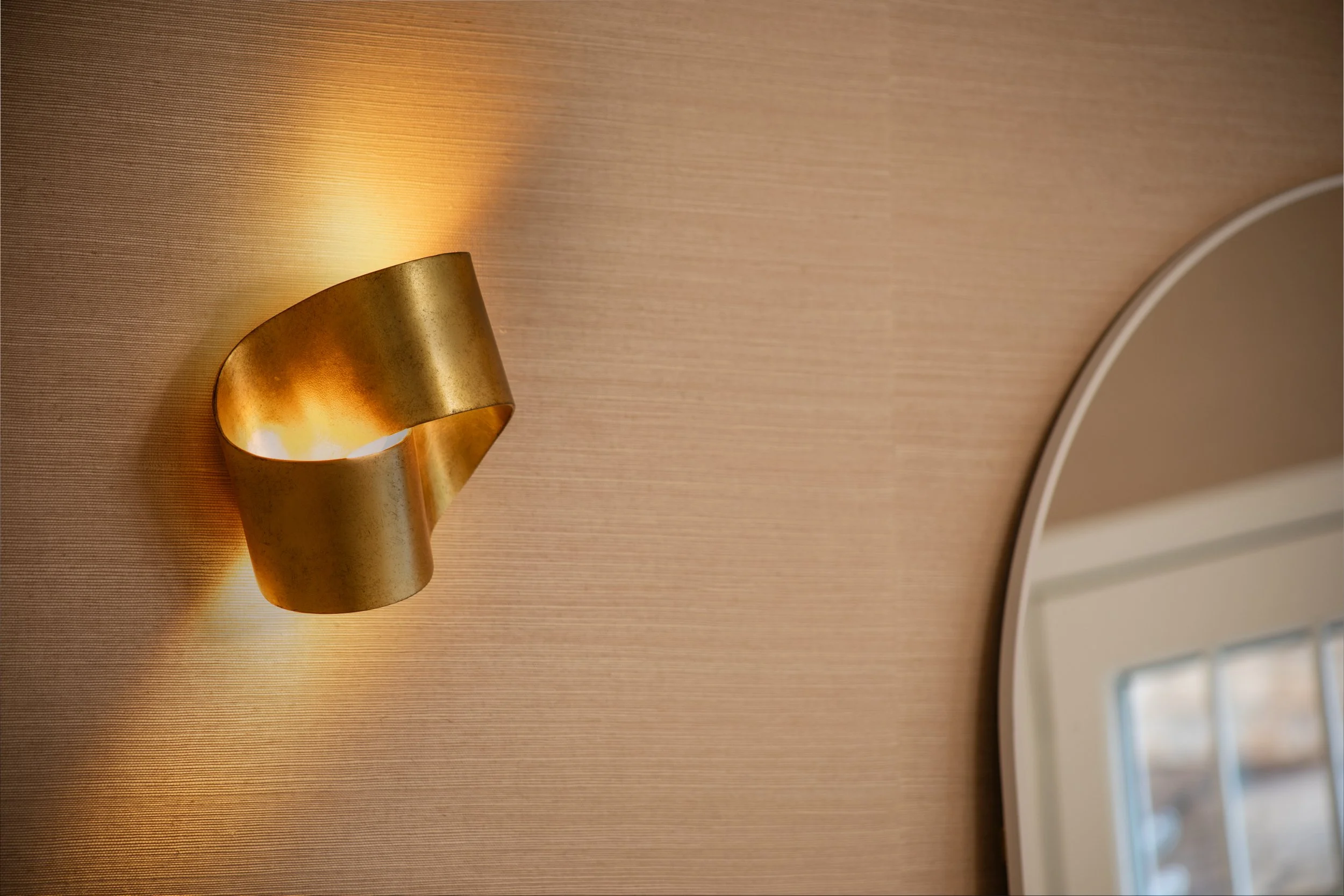 Golden wall-mounted lamp casting warm light on a textured beige wall, with part of a round mirror visible on the right.