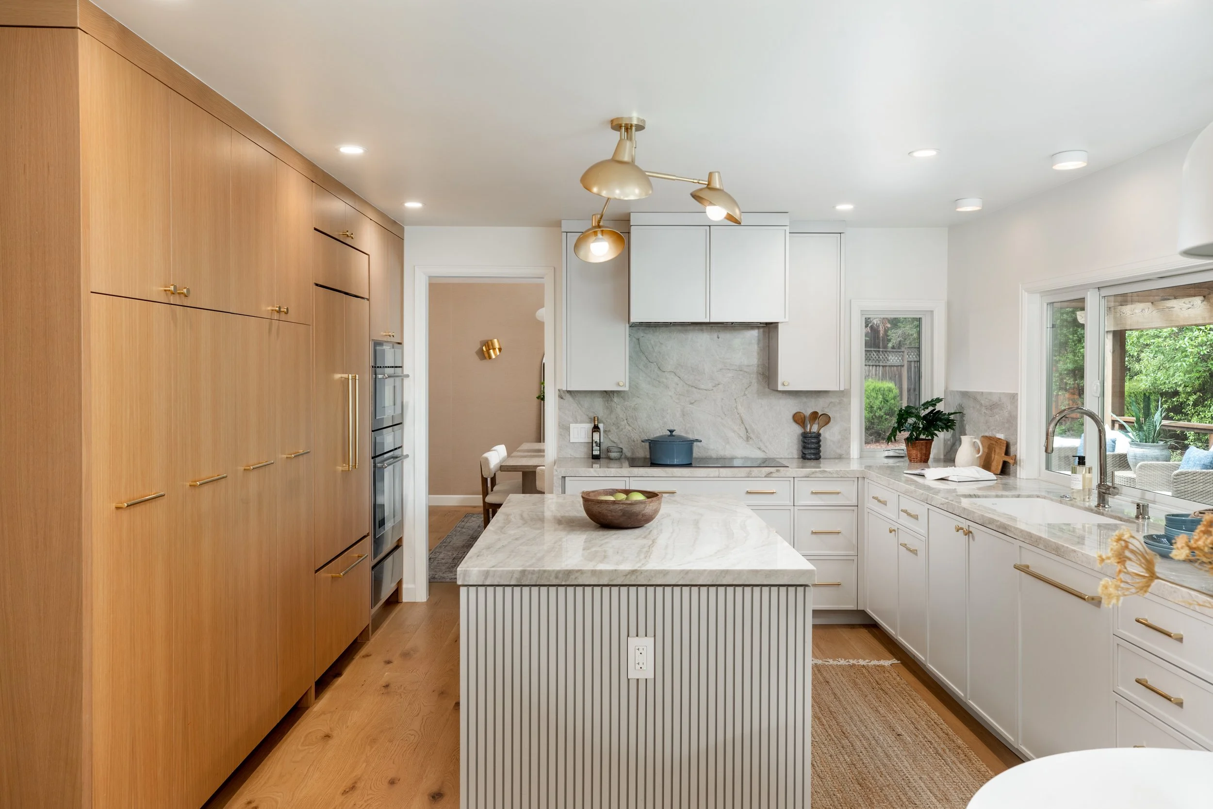 A modern kitchen with white cabinets, a marble island, wooden ceiling-high cabinets with gold handles, and large windows looking into a garden.