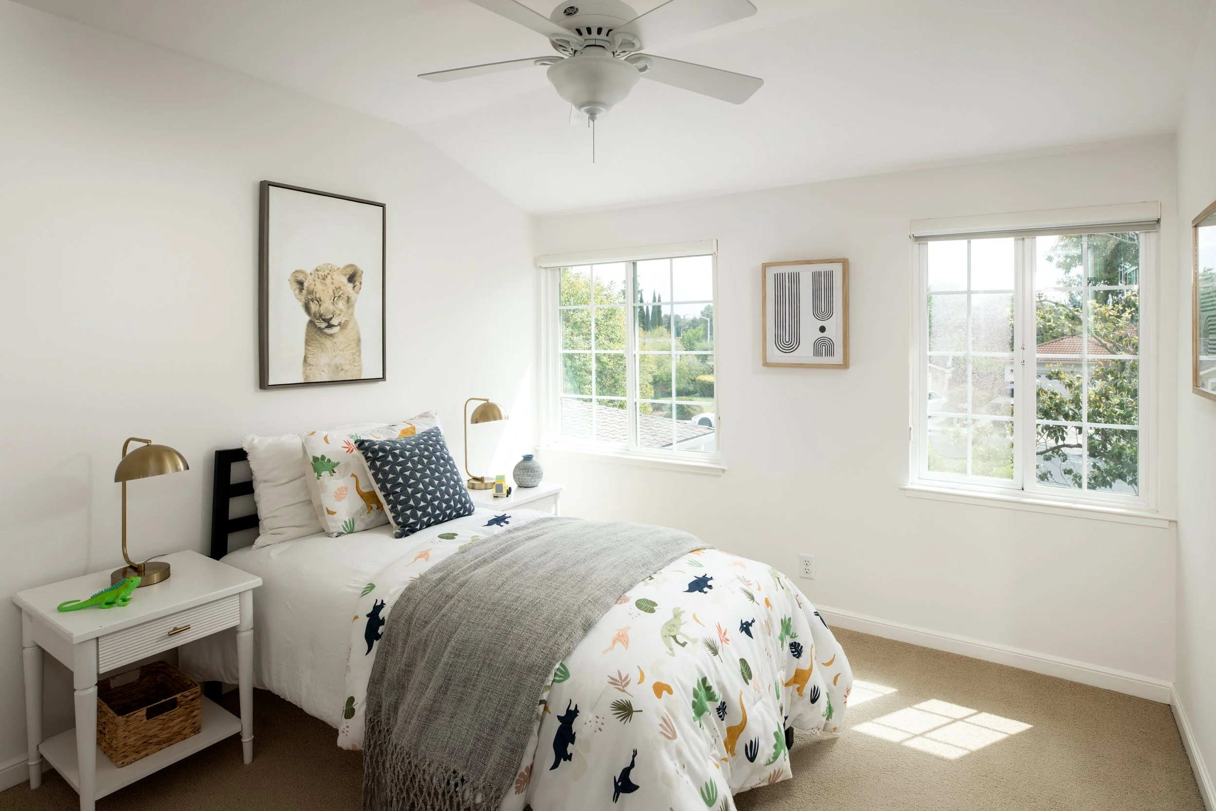 A bedroom with white walls and large windows showing trees outside. A bed with a dinosaur-themed comforter and decorative pillows is against the wall. On the bedside table, there are two brass-colored lamps, a small green dinosaur figurine, and a gra