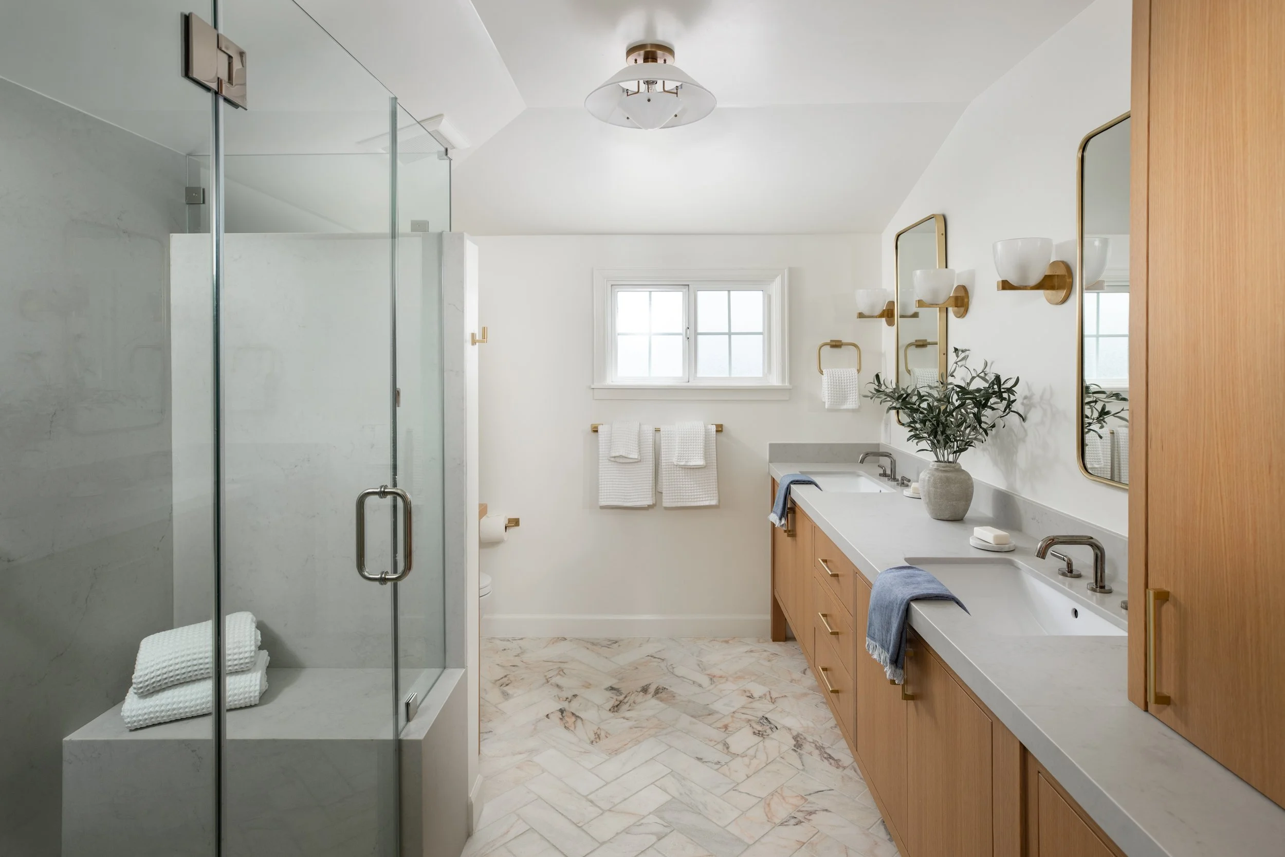 A modern bathroom with a glass-enclosed shower, a wooden vanity with a white countertop, two mirrors, white towels, a window, a potted plant, and marble flooring.