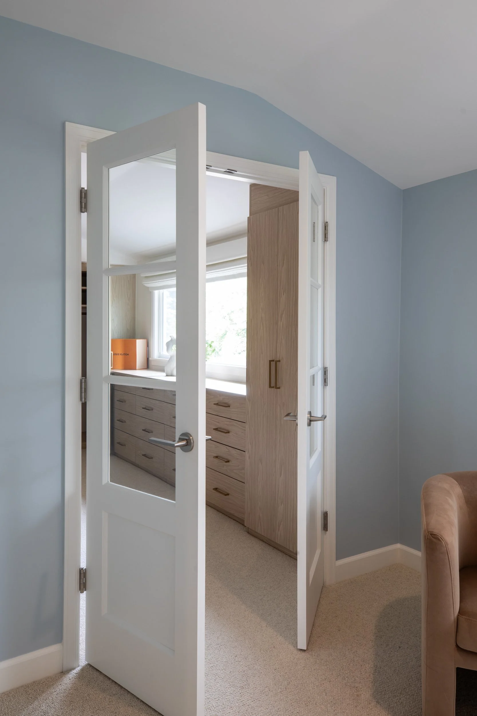 A white half-glass door is open, revealing a room with a light wood storage cabinet and a window with white blinds. A brown armchair is partially visible on the right, and light blue painted walls and beige carpeted flooring are seen.