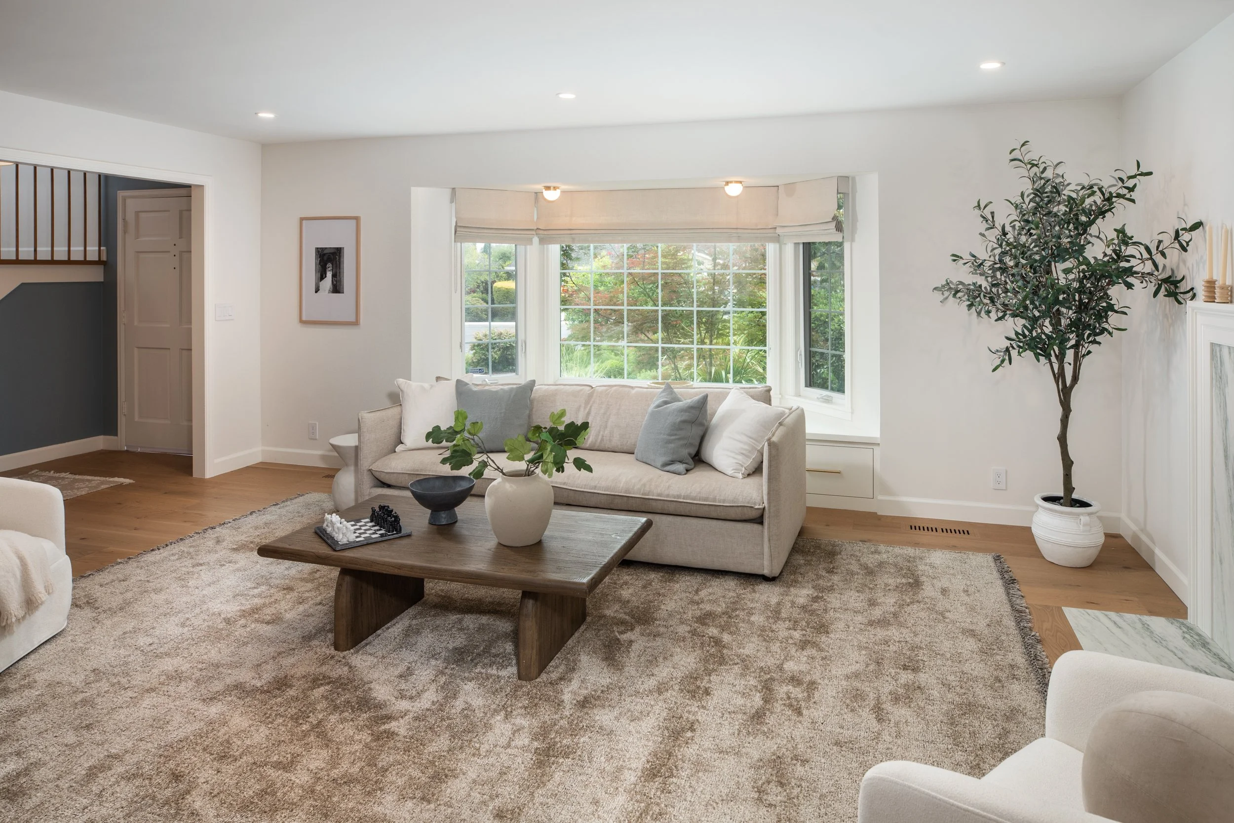 Living room with cream-colored sofa, large bay window with beige roman shades, wooden coffee table with a potted plant, framed black-and-white photo on the wall, and a tall leafy plant in a white pot.