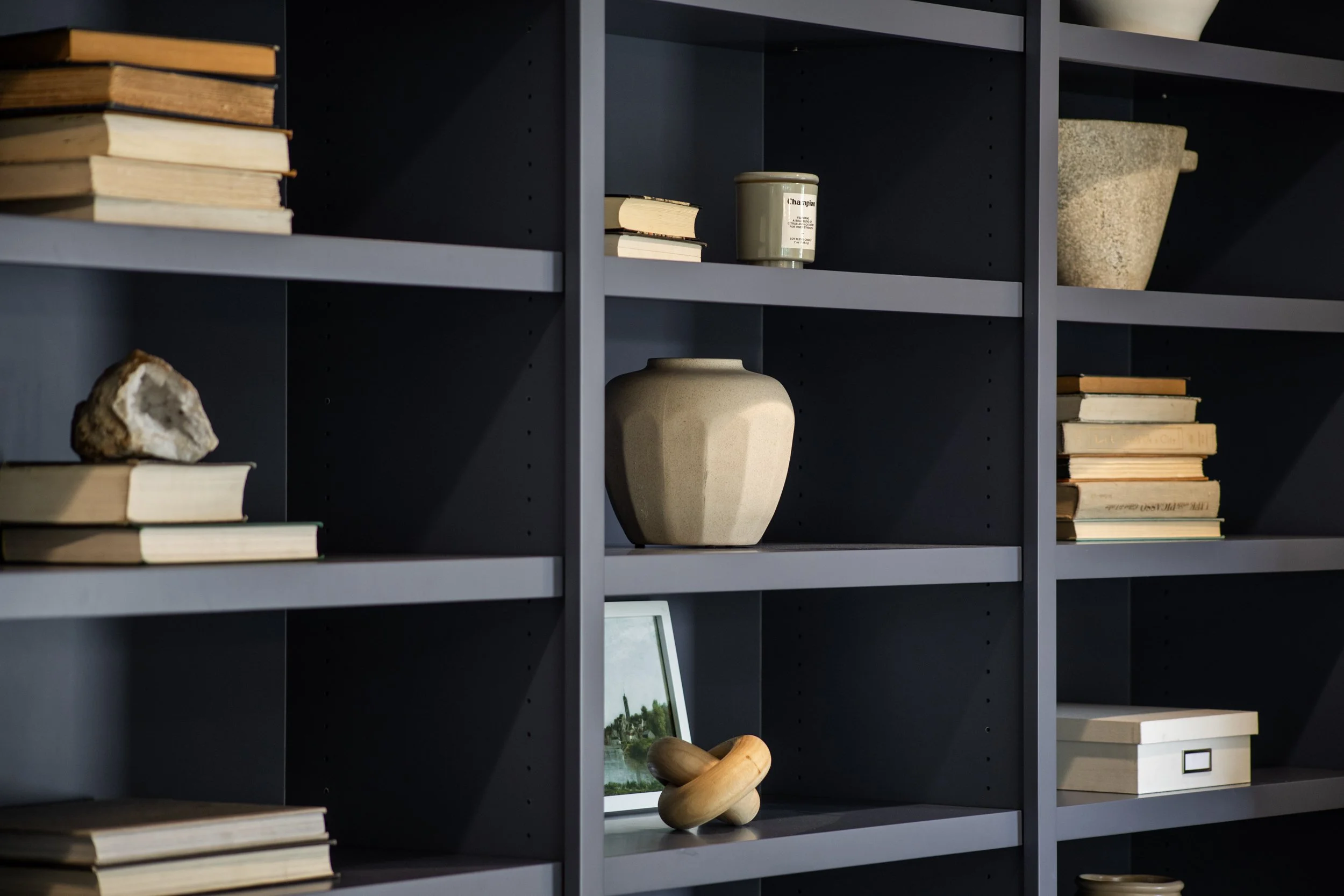 Dark gray bookshelf with various decorative items including stacks of books, ceramic jars, a framed photograph of a lighthouse, a stone sculpture, and a small wooden sculpture.