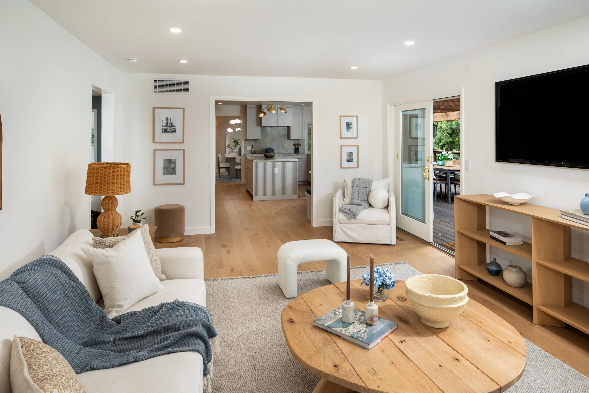 Living room with white walls, wooden furniture, a blue sofa with white and beige pillows, a coffee table with candles and books, a bookshelf, a white armchair with a throw, and an open view to a kitchen and patio with trees outside.