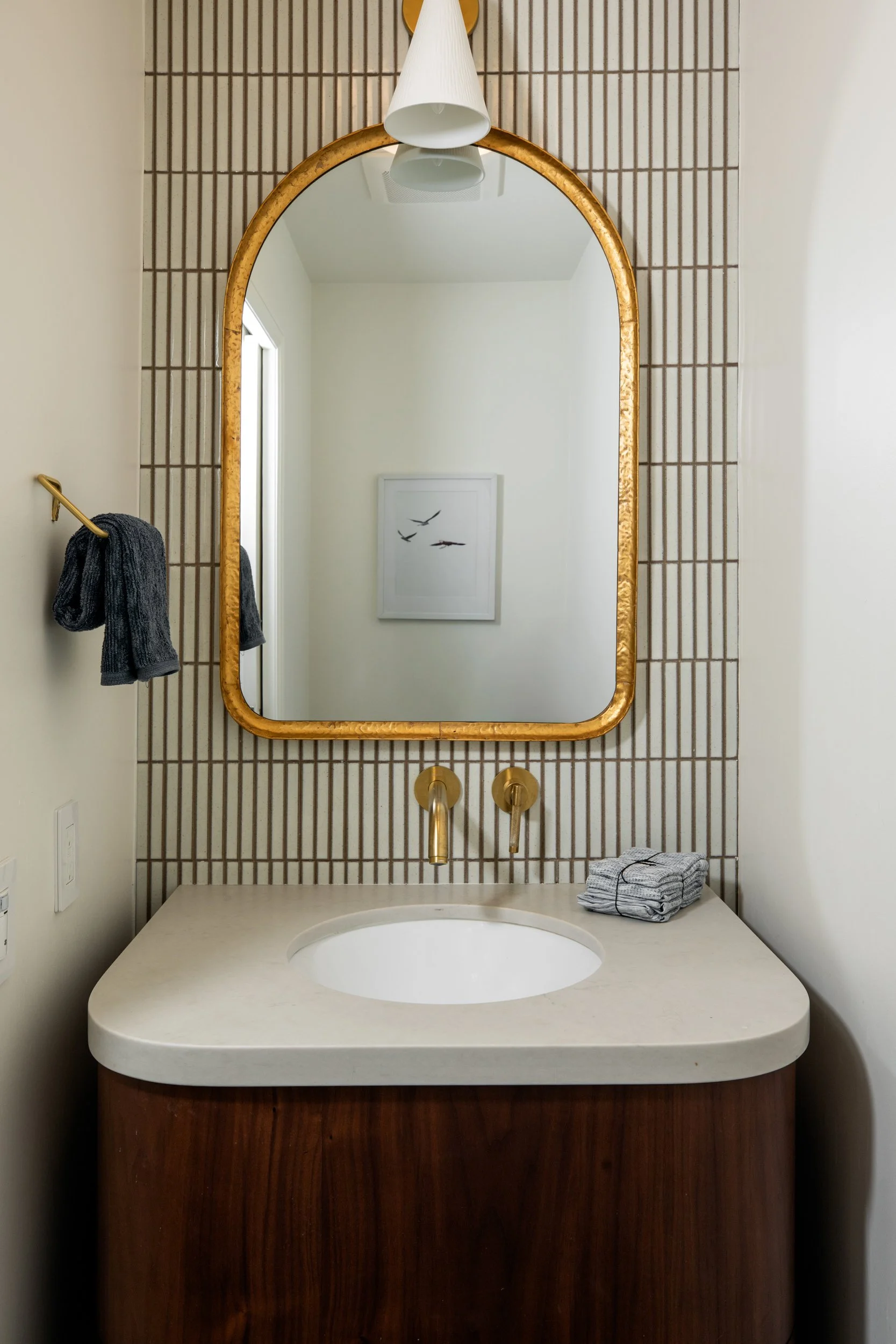A bathroom vanity with a round opening sink, brass faucet, gold framed mirror, and a white countertop, with a striped tile wall and a framed bird print in the background.