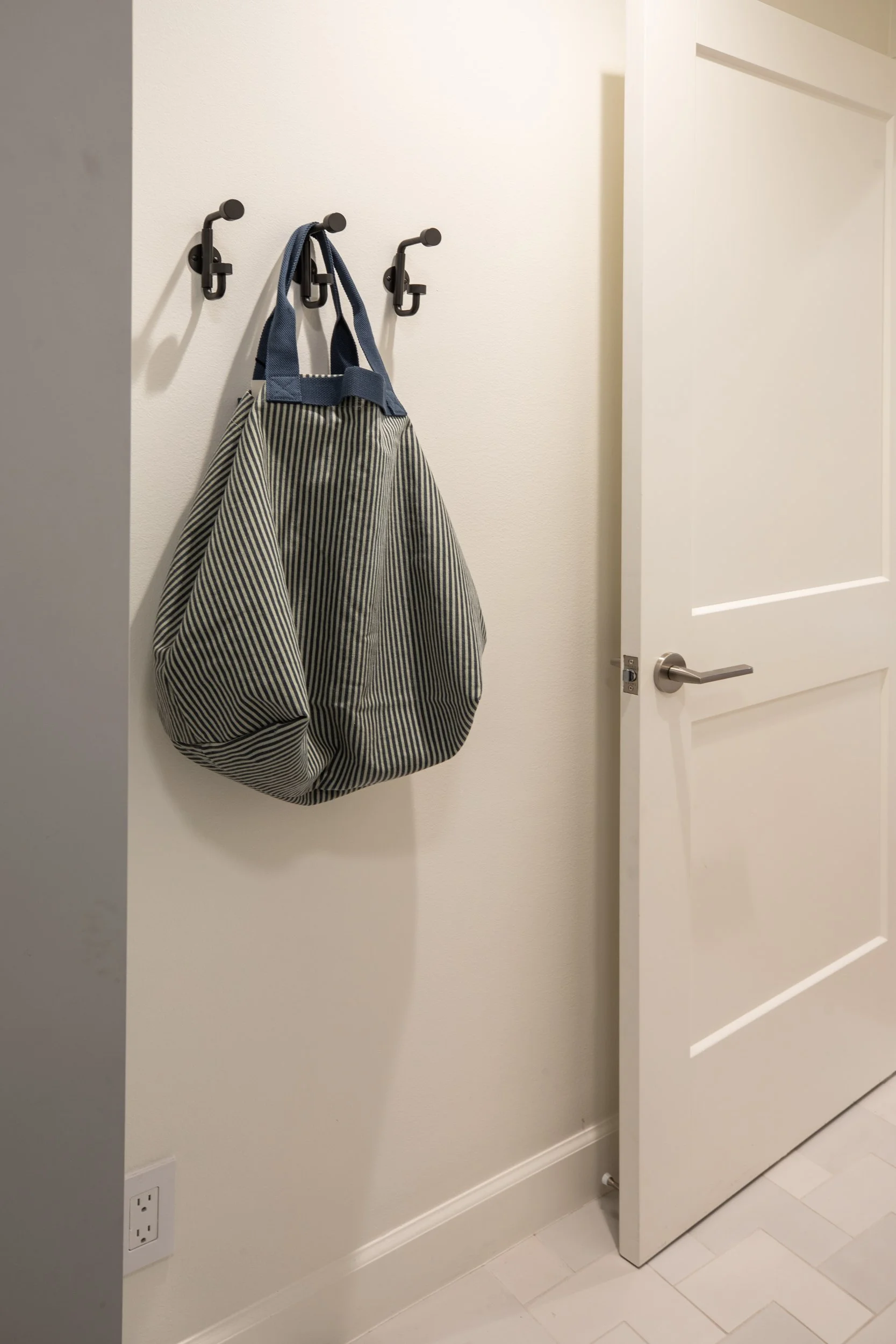 A hallway with hooks on the wall, one holding a striped bag with a blue strap. There is an open white door with a silver handle. The wall is plain and painted light beige, with a power outlet near the floor.