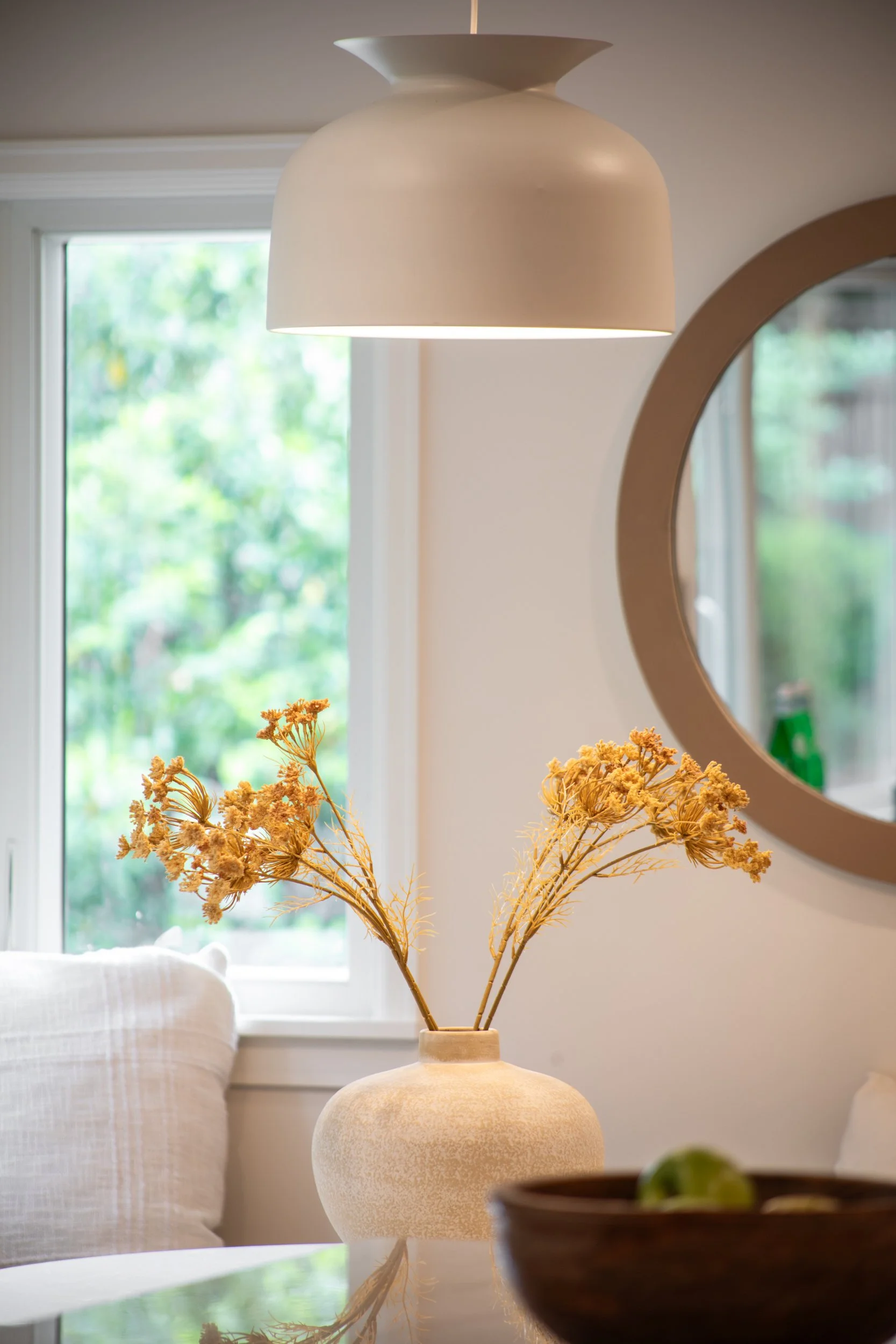 Close-up of a beige vase with dried yellow flowers on a reflective surface, with a background of a window and interior mirror.