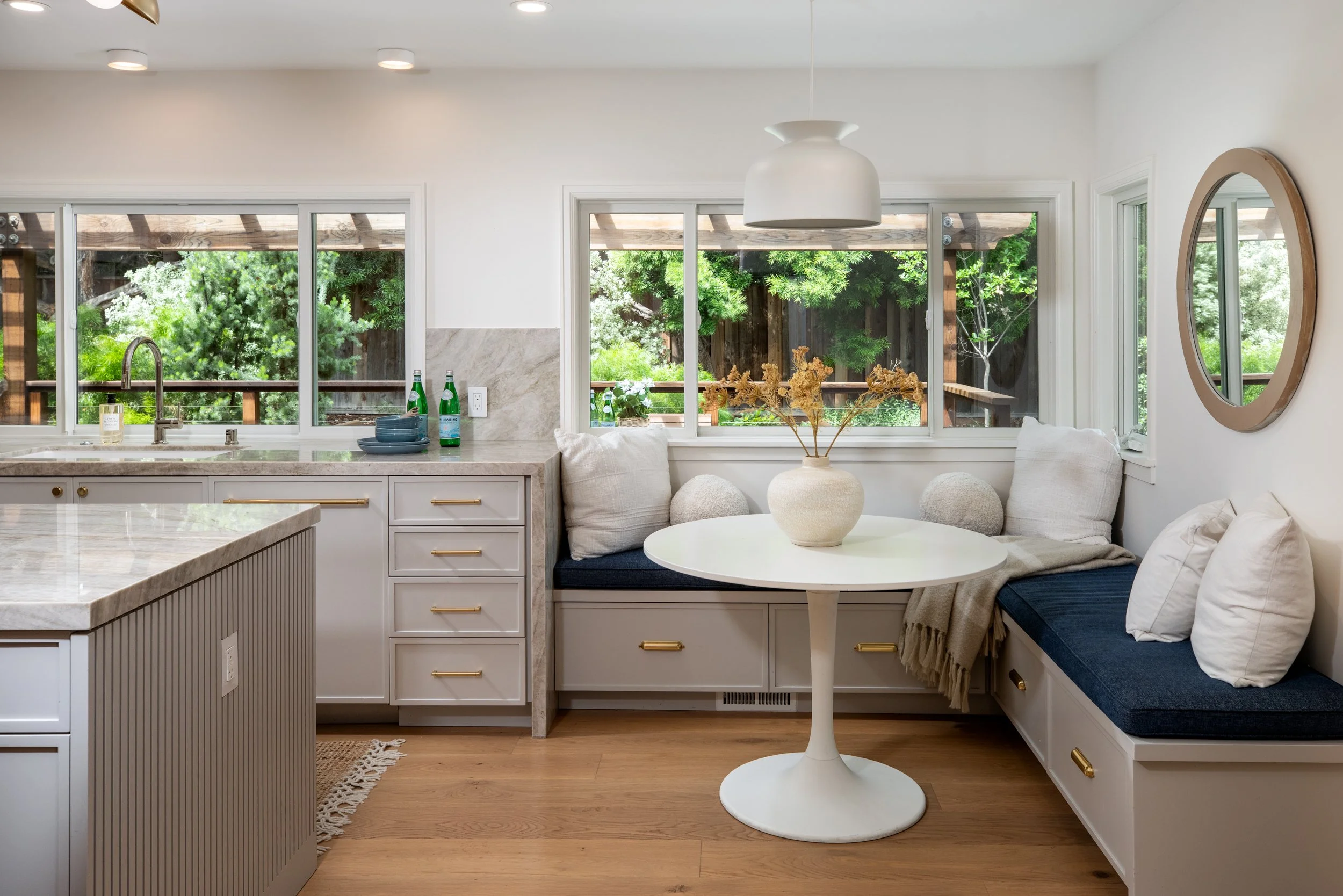 A bright kitchen with large windows looking out onto a lush green outdoor scene. The kitchen has white cabinets, a marble countertop, and a round white table with a floral centerpiece. There are two green glass bottles and some dishes on the countert