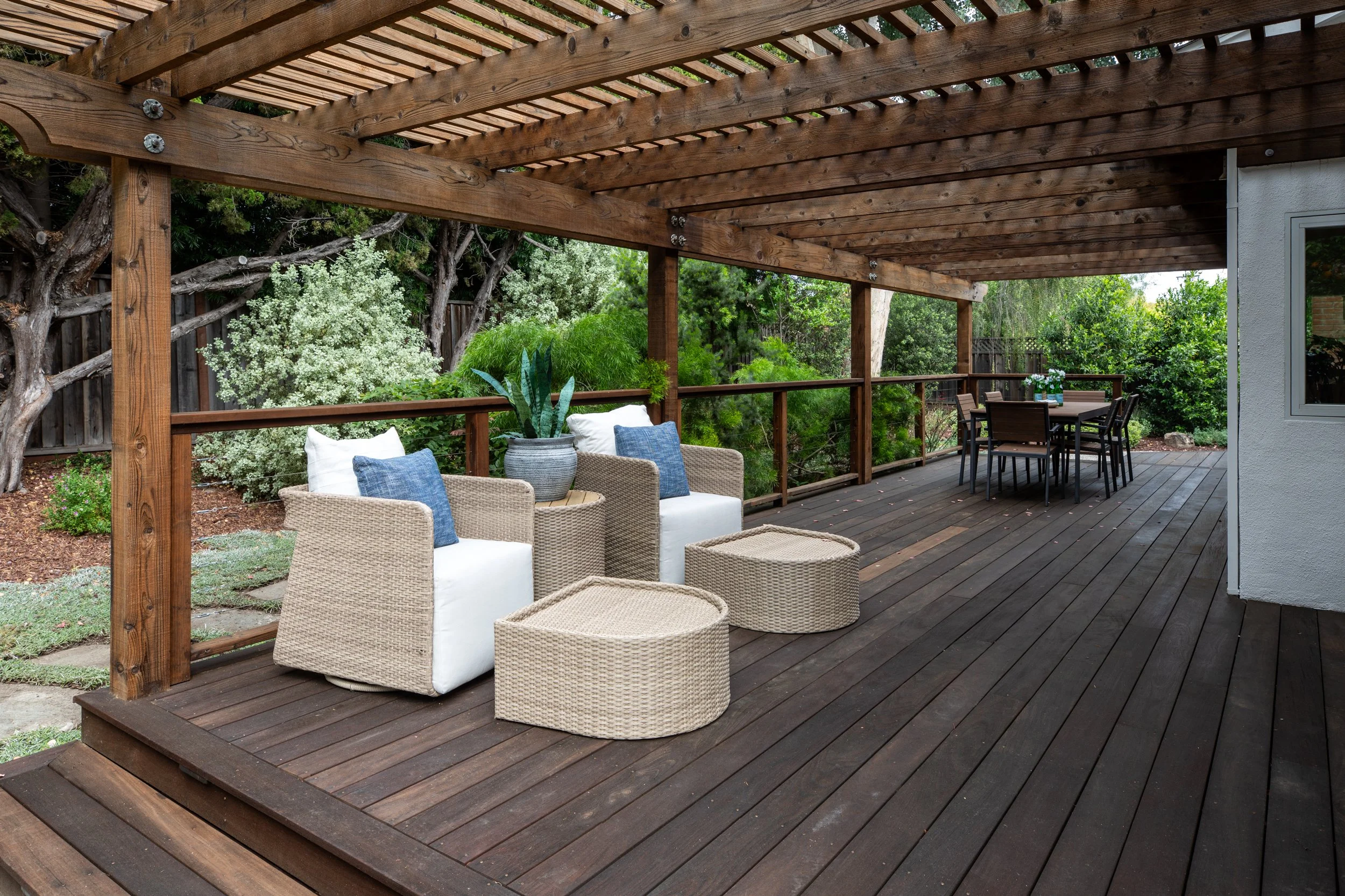 Wooden patio with seating area, including wicker chairs with white cushions and blue pillows, a potted plant, and an outdoor dining table with chairs, surrounded by lush greenery and trees.