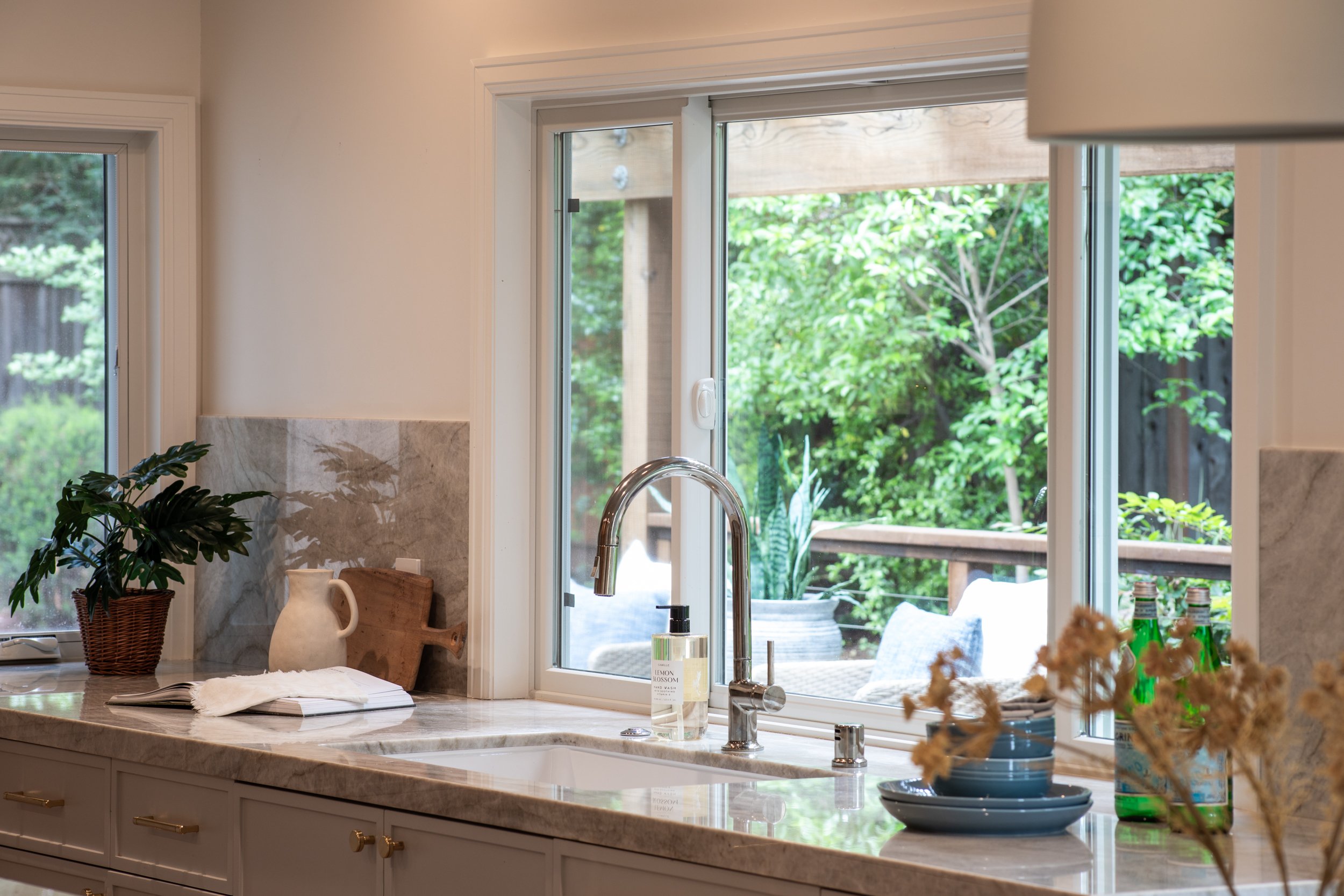 Kitchen countertop with a potted plant, a white pitcher, a cutting board, an open book, and a soap dispenser, with a large window showing a green backyard outside.