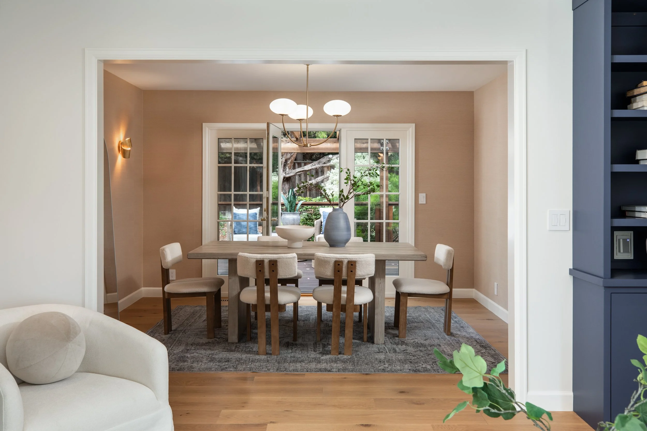 A dining room with French doors leading to a backyard, a wooden dining table with six chairs, and a modern chandelier hanging above the table. The room has beige walls and a dark blue bookshelf on the right.