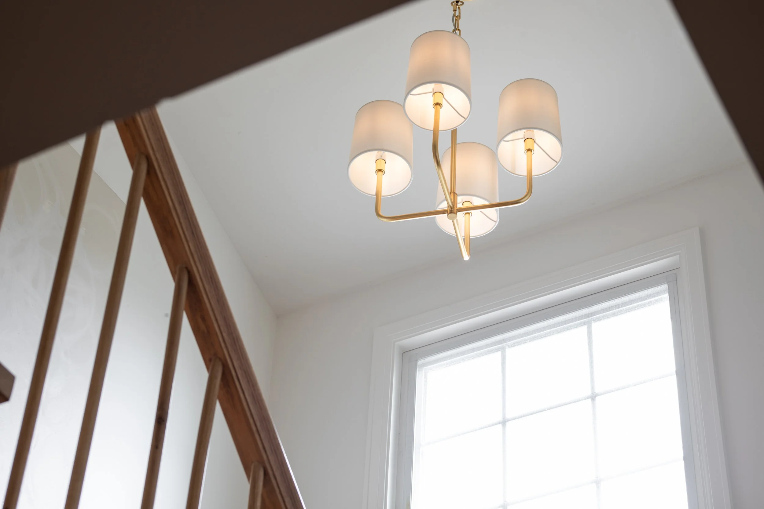Ceiling light fixture with four frosted glass lampshades and gold metal accents, viewed from below through a staircase railing, with a window in the background.
