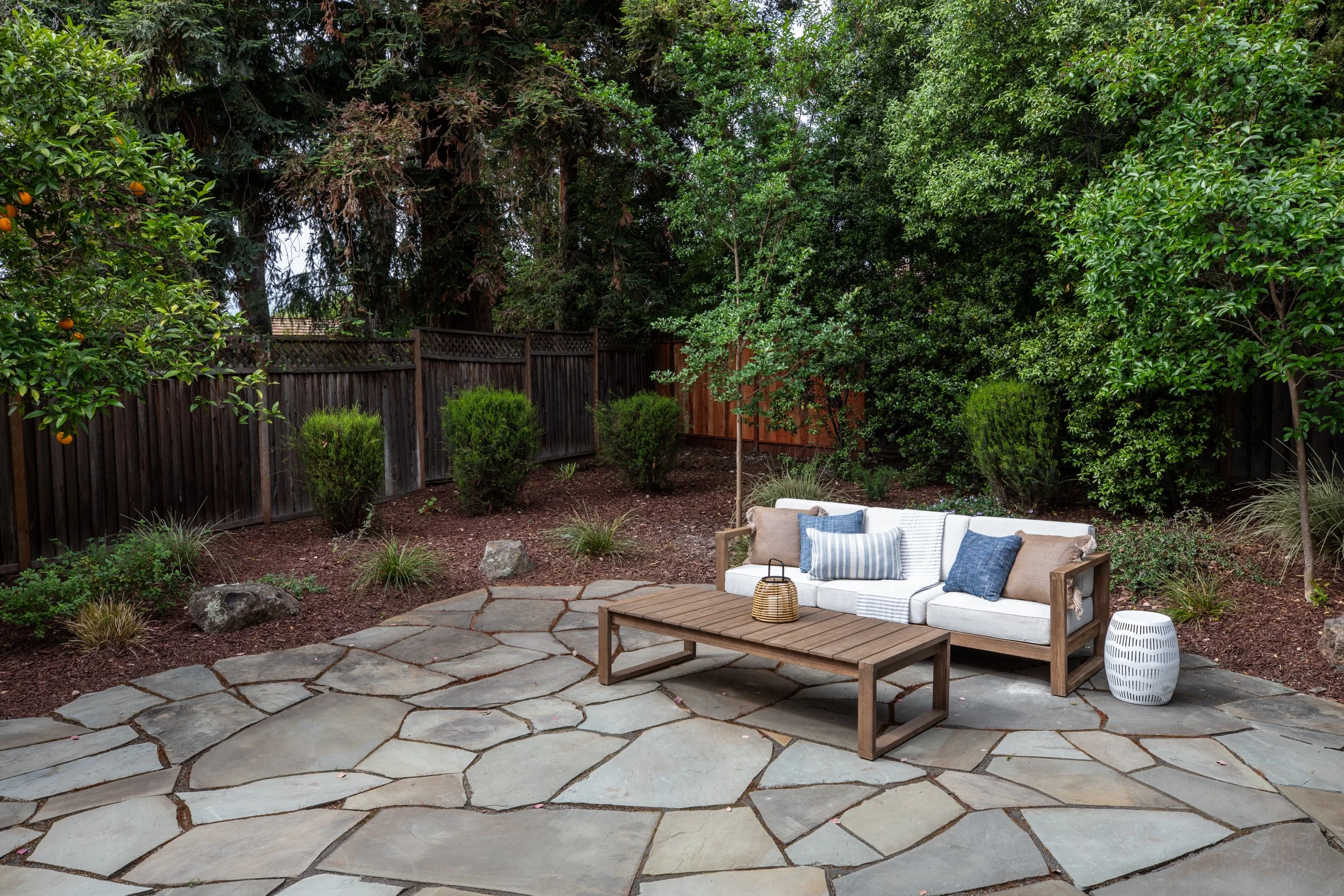 A backyard with a stone patio, a white outdoor sofa with pillows, a wooden coffee table, and a white side table, surrounded by lush green trees and bushes, with a wooden fence in the background.