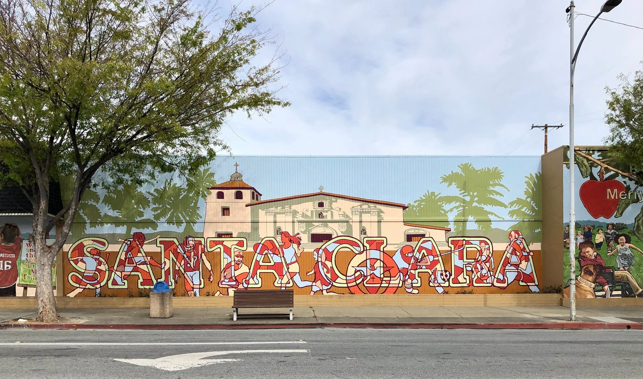 Colorful mural art on a wall depicting a church and people playing sports such as volleyball and soccer, with trees and a cloudy sky above, located next to a street with a bench and a trash can.