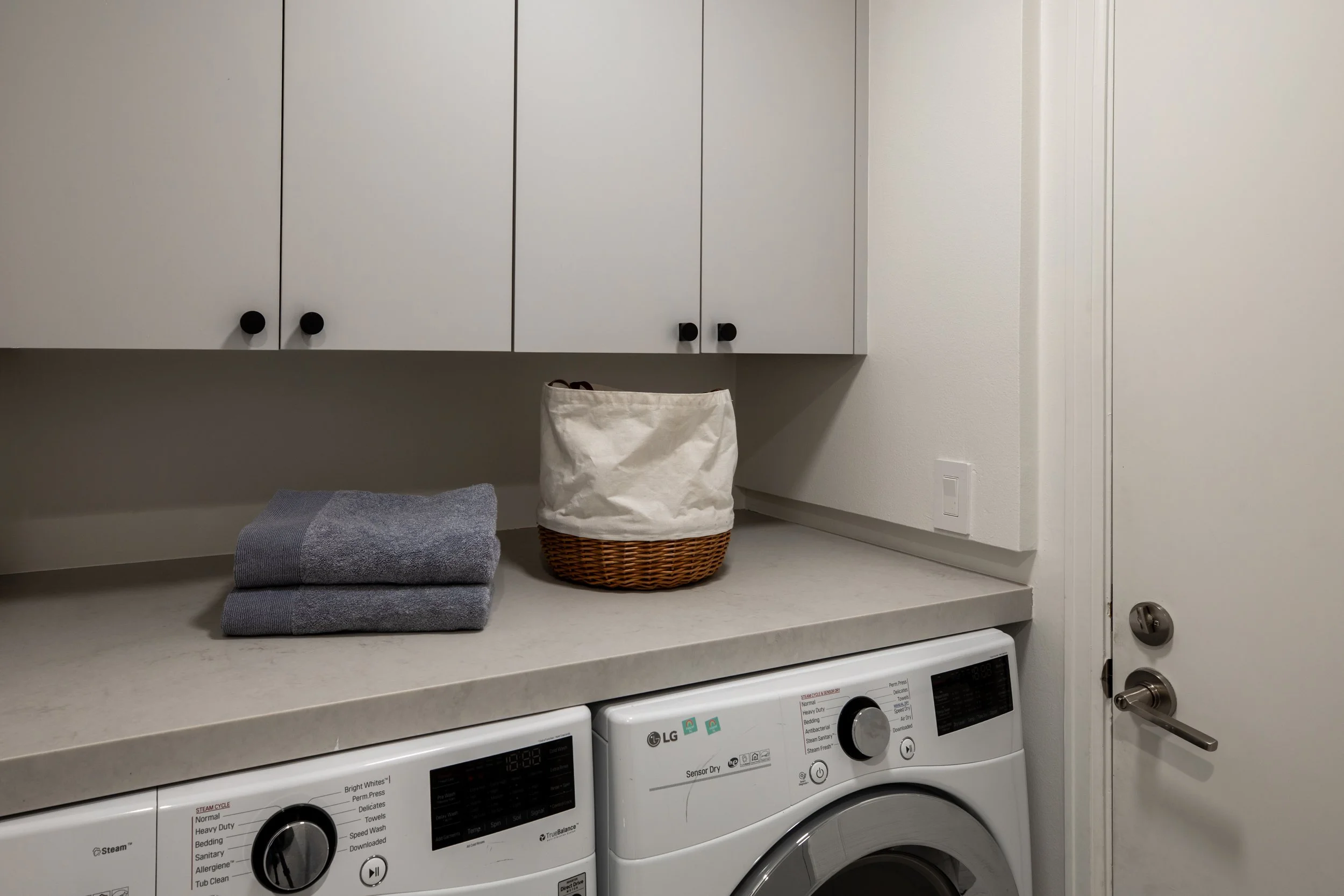 A laundry area with four white cabinets on the wall, a gray folded towel, a basket lined with a cloth bag, and a white LG washing machine.