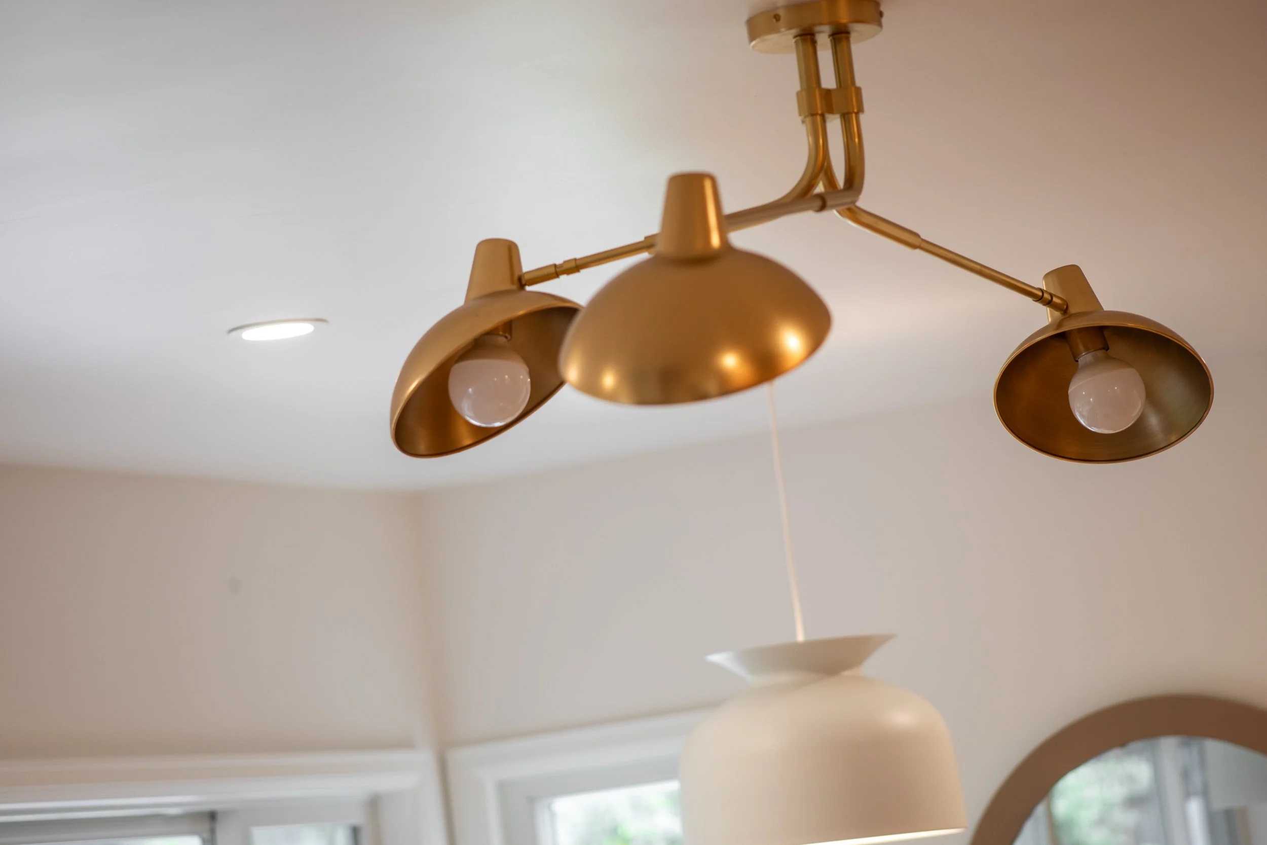 Gold-colored ceiling light fixture with multiple adjustable arms and white bulbs, mounted on a white ceiling in a room with windows and light-colored walls.