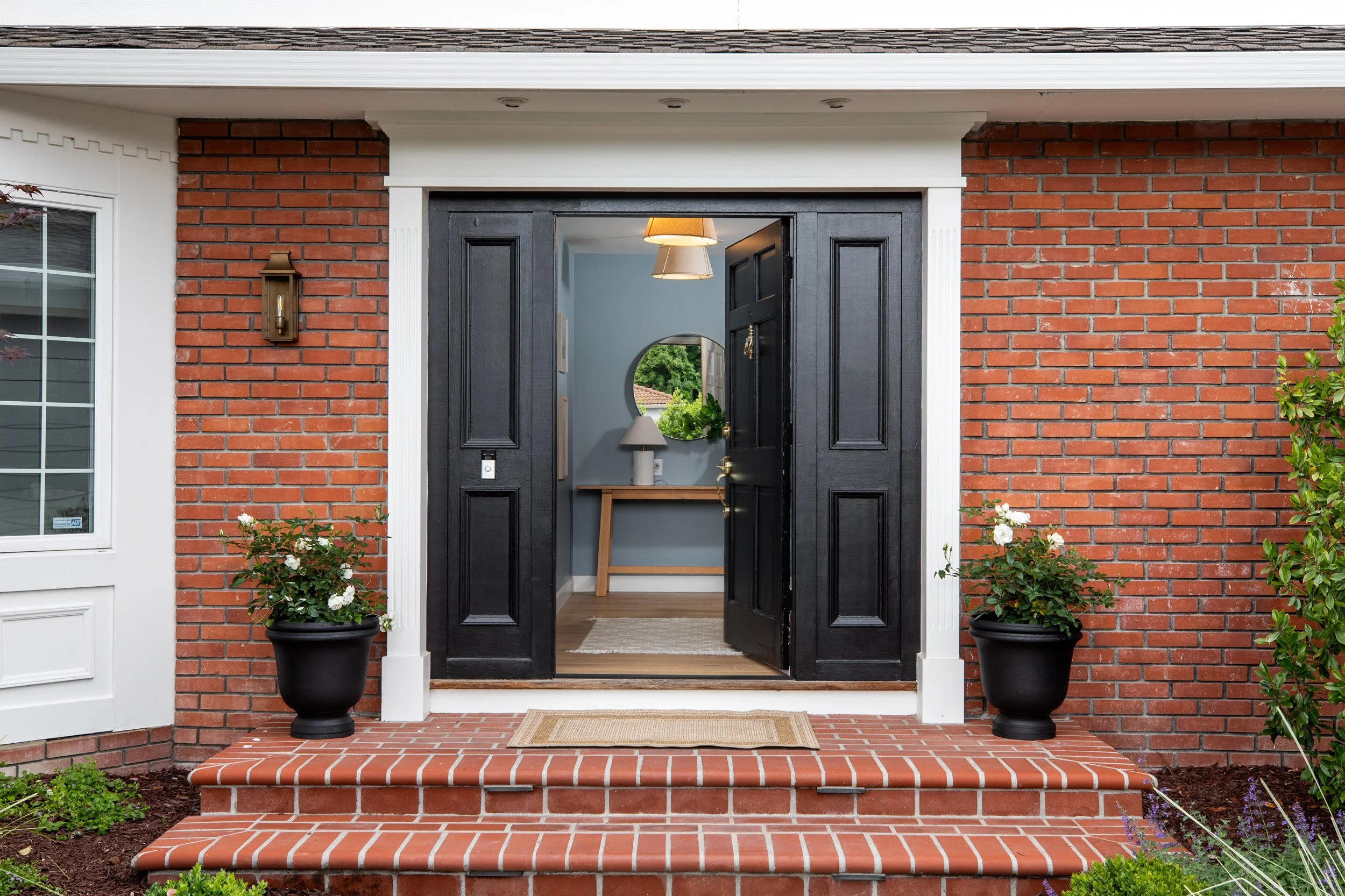 The front entrance of a house with brick steps, black double doors, two potted plants on each side, and a verandah with light fixtures and a mirror inside visible through the open doors.